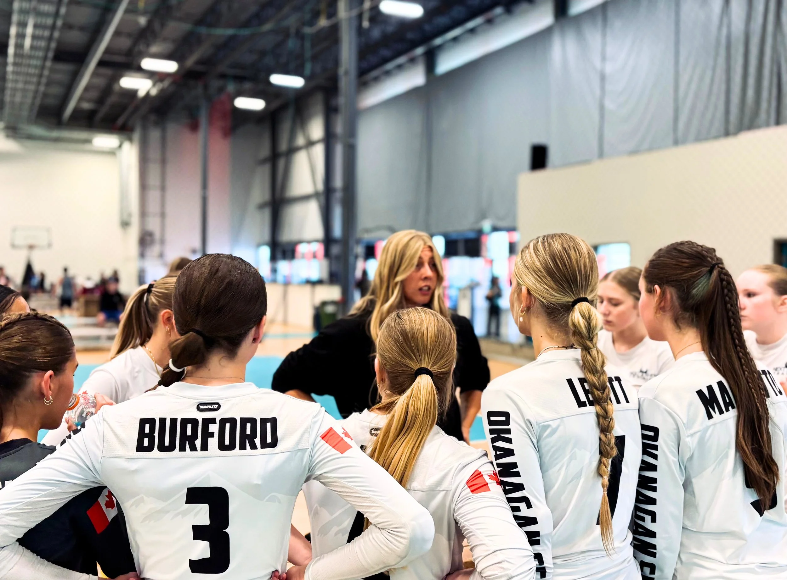 A group of female volleyball players in white uniforms gathered in huddle inside a gymnasium, with some showing their last names and numbers on the back of their jerseys.