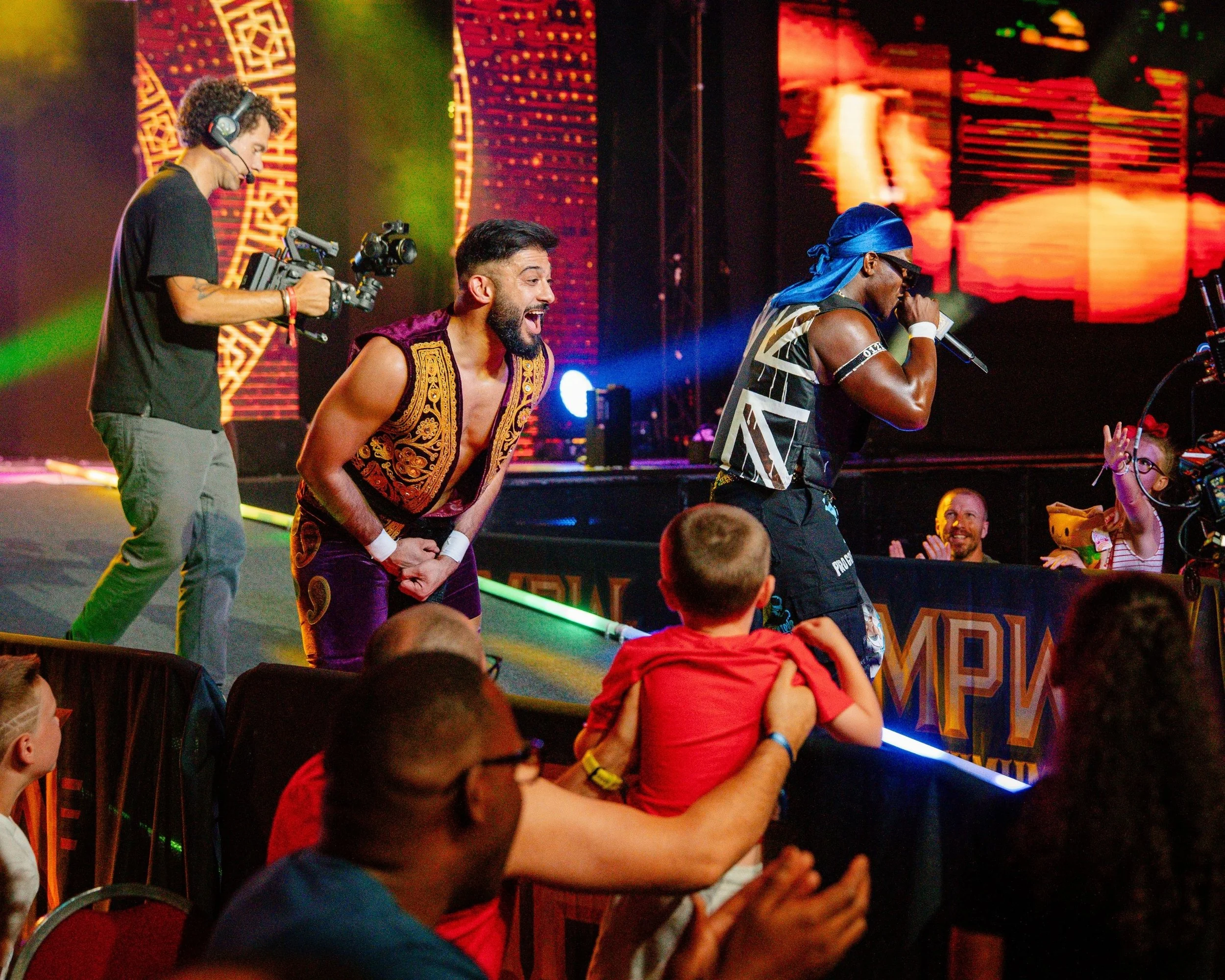 A vibrant stage performance featuring a singer with a blue bandana and sunglasses, another performer smiling and wearing a sleeveless embroidered vest, and a cameraman filming the scene. Audience members interact in front of the stage with colorful backdrop screens.