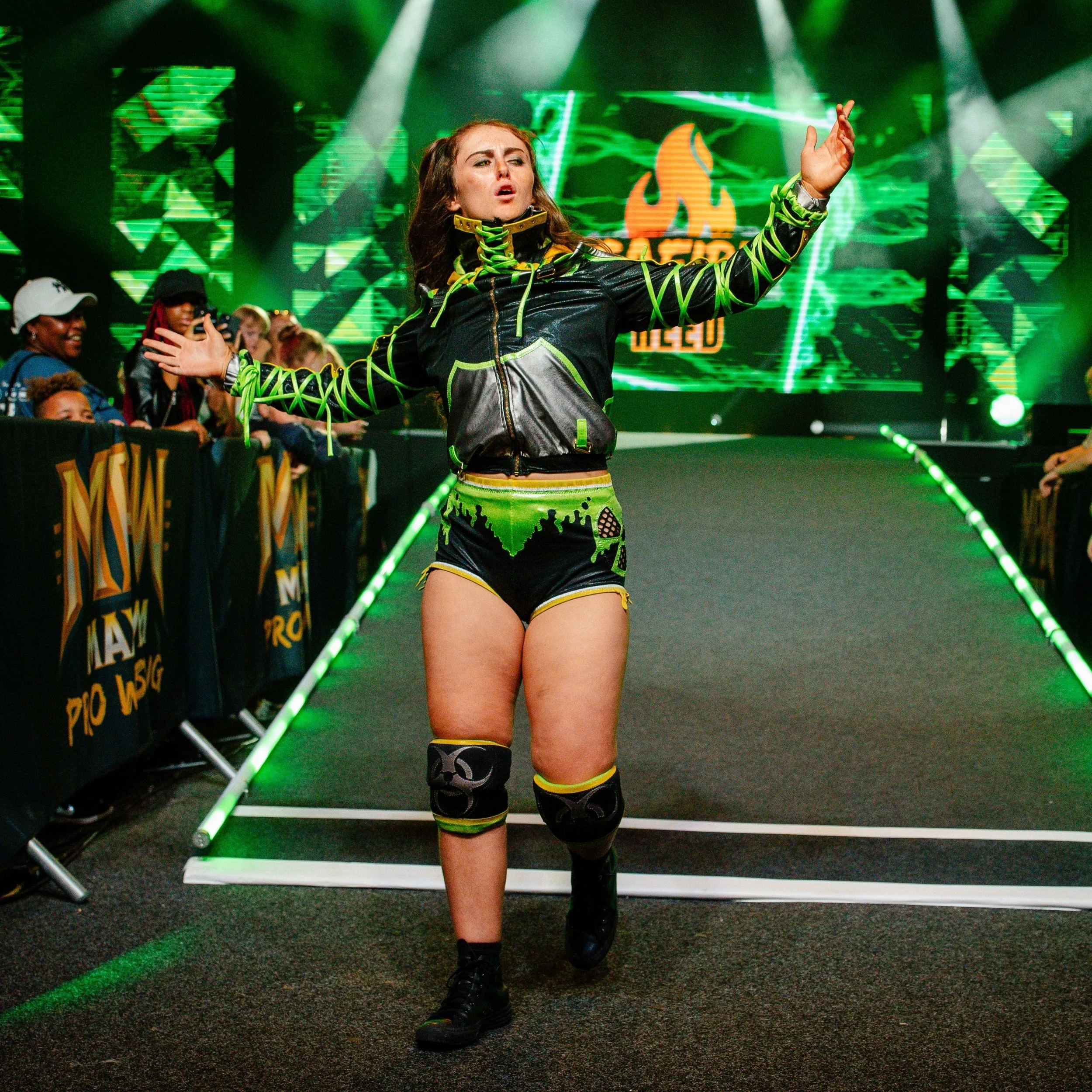 Female wrestler posing backstage at wrestling event, with green and black attire, tattoos, and a focused expression, illuminated by green stage lighting and digital background.