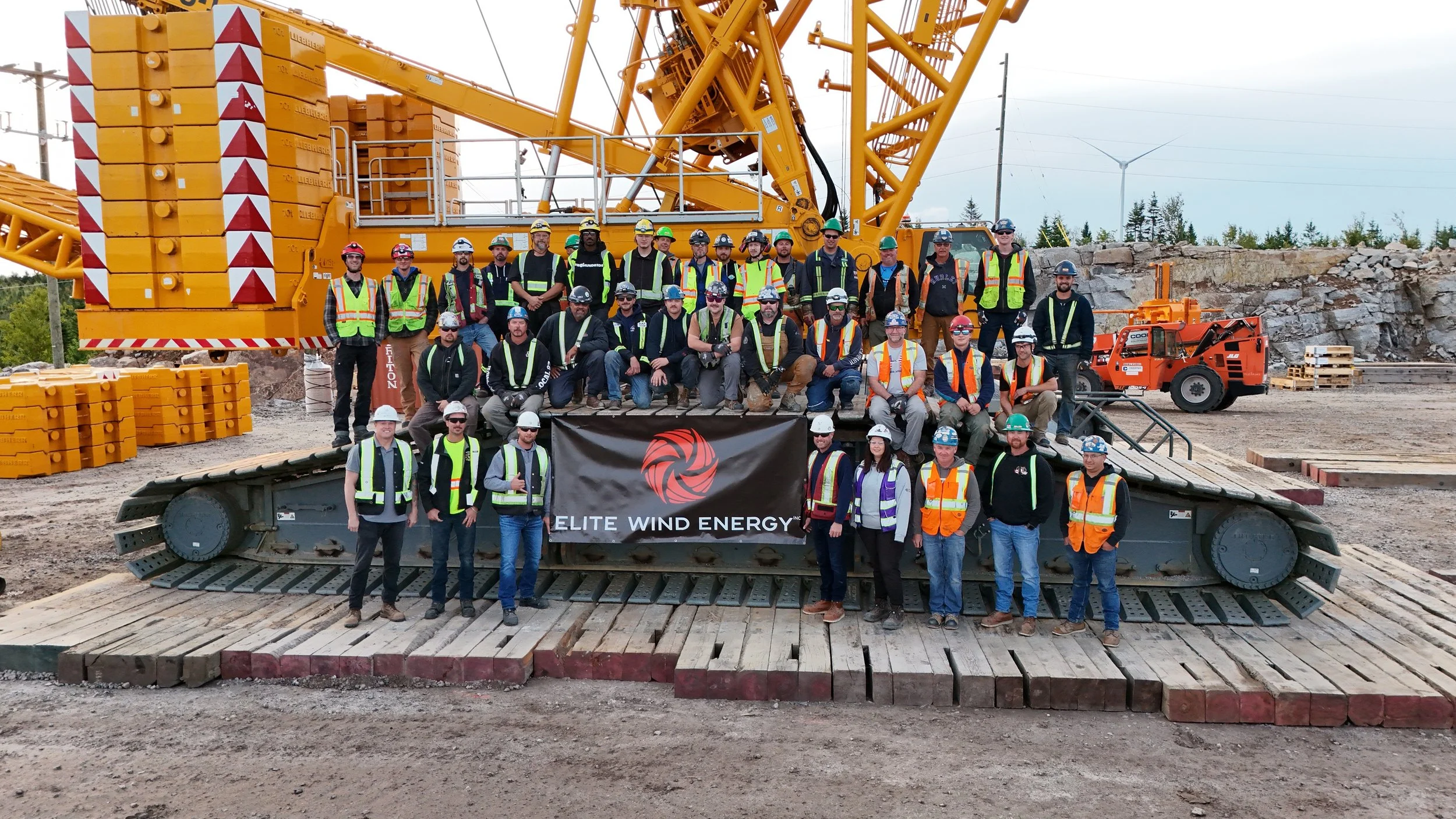 Group of construction workers and engineers wearing safety gear, including vests and helmets, posing on a large tracked vehicle with a yellow crane in the background at a wind energy construction site. A black banner with red logo and the text 'ELITE WIND ENERGY' is displayed in front.
