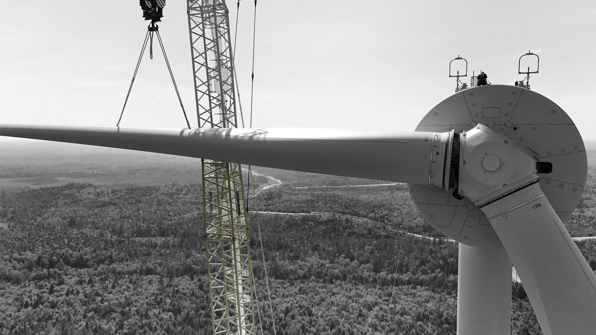 Black and white photo of a wind turbine being assembled with a construction crane next to the tower, high above a forested landscape.