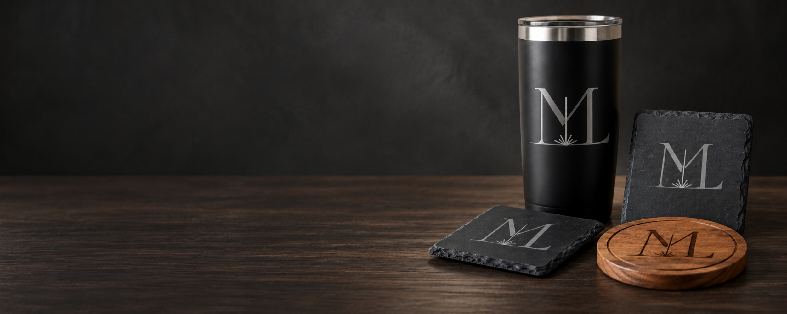 A black travel tumbler, two black slate coasters, and a round wooden coaster, all with a white or black logo featuring the letters 'ML' and an abstract design, on a wooden surface against a dark background.