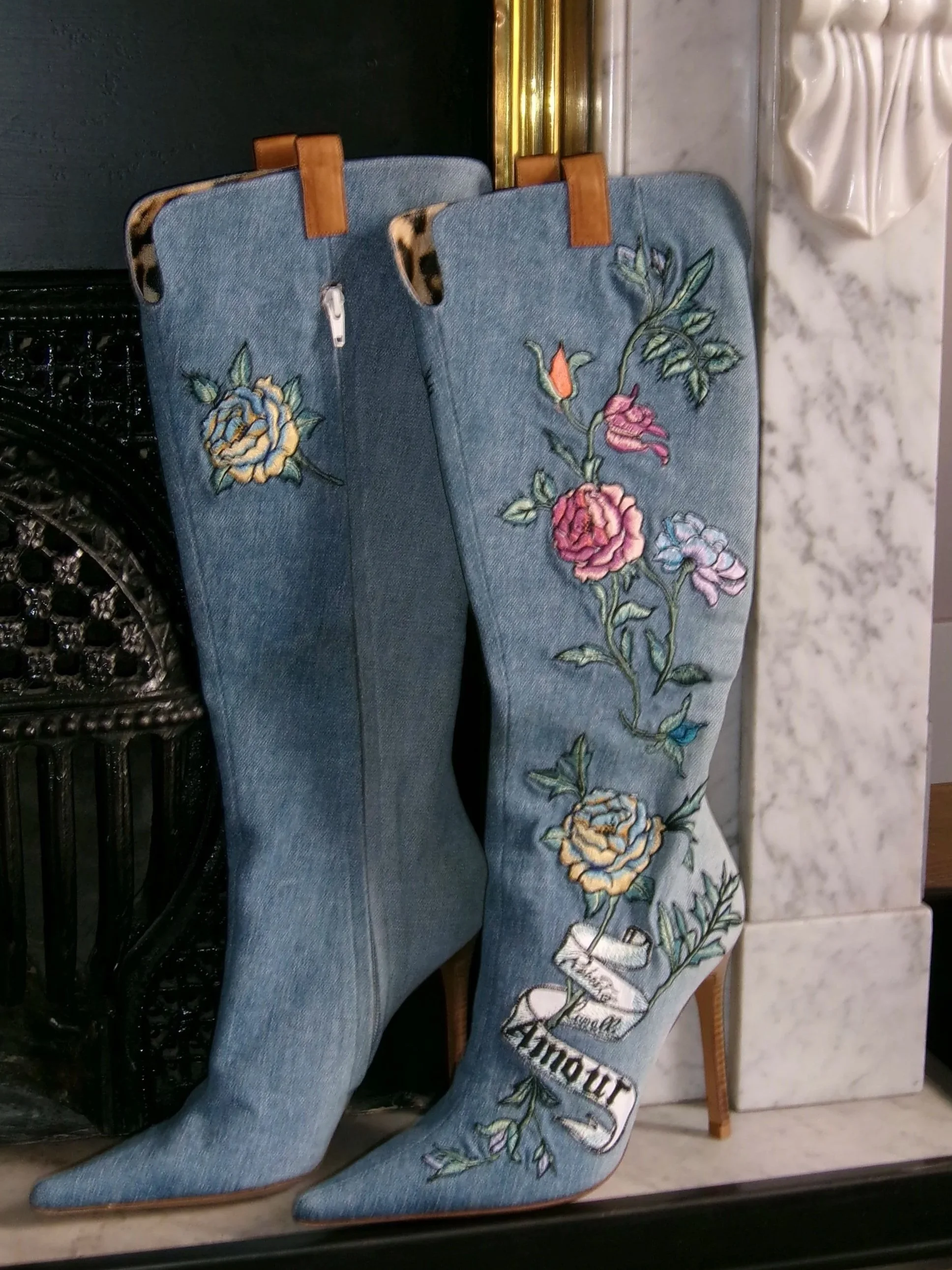 Pair of embroidered denim cowboy boots with floral designs, displayed on a shelf in a store.