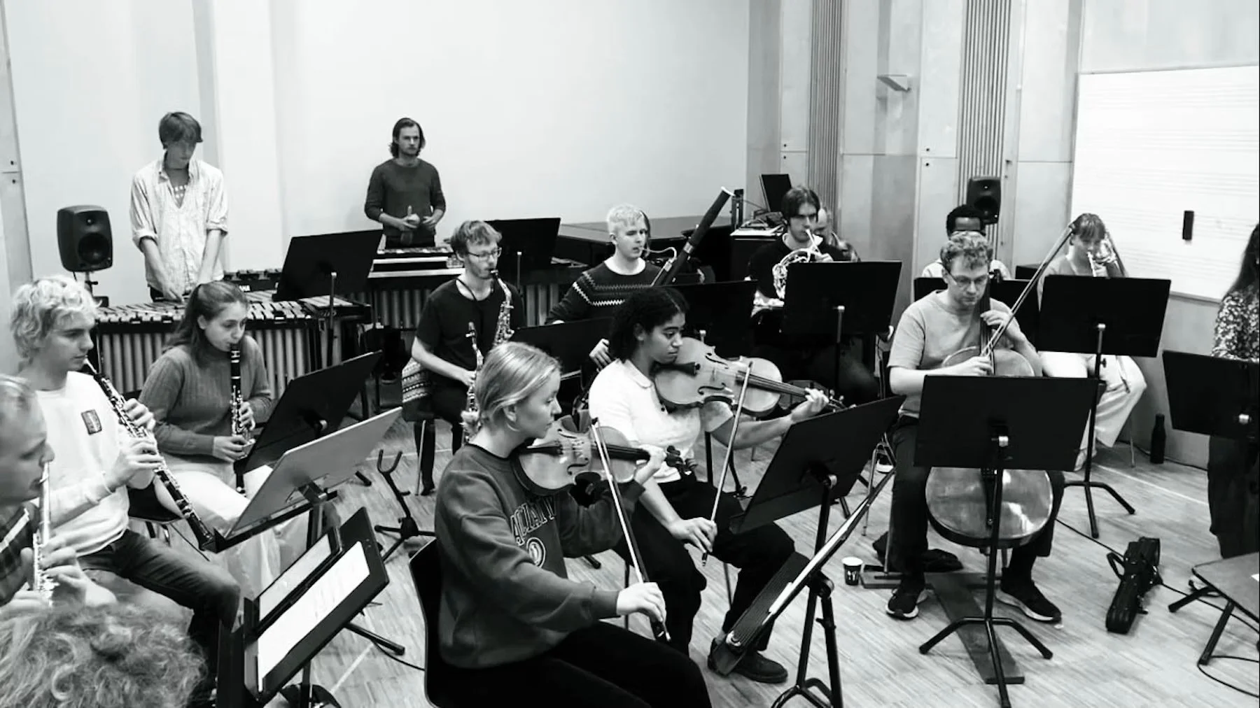 A group of musicians rehearsing in a music studio, playing various instruments including violins, clarinets, and a cello, with a conductor or instructor standing on the right.