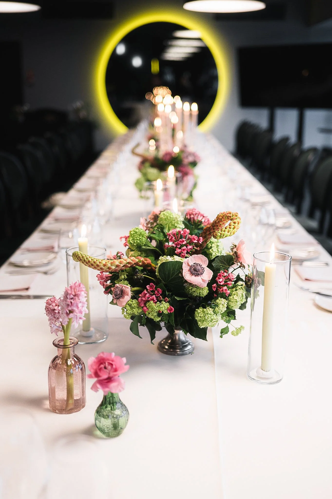 A long dining table decorated with pink and green floral arrangements in small vases and lit candles, with black chairs and a dark background featuring a large circular yellow light feature.