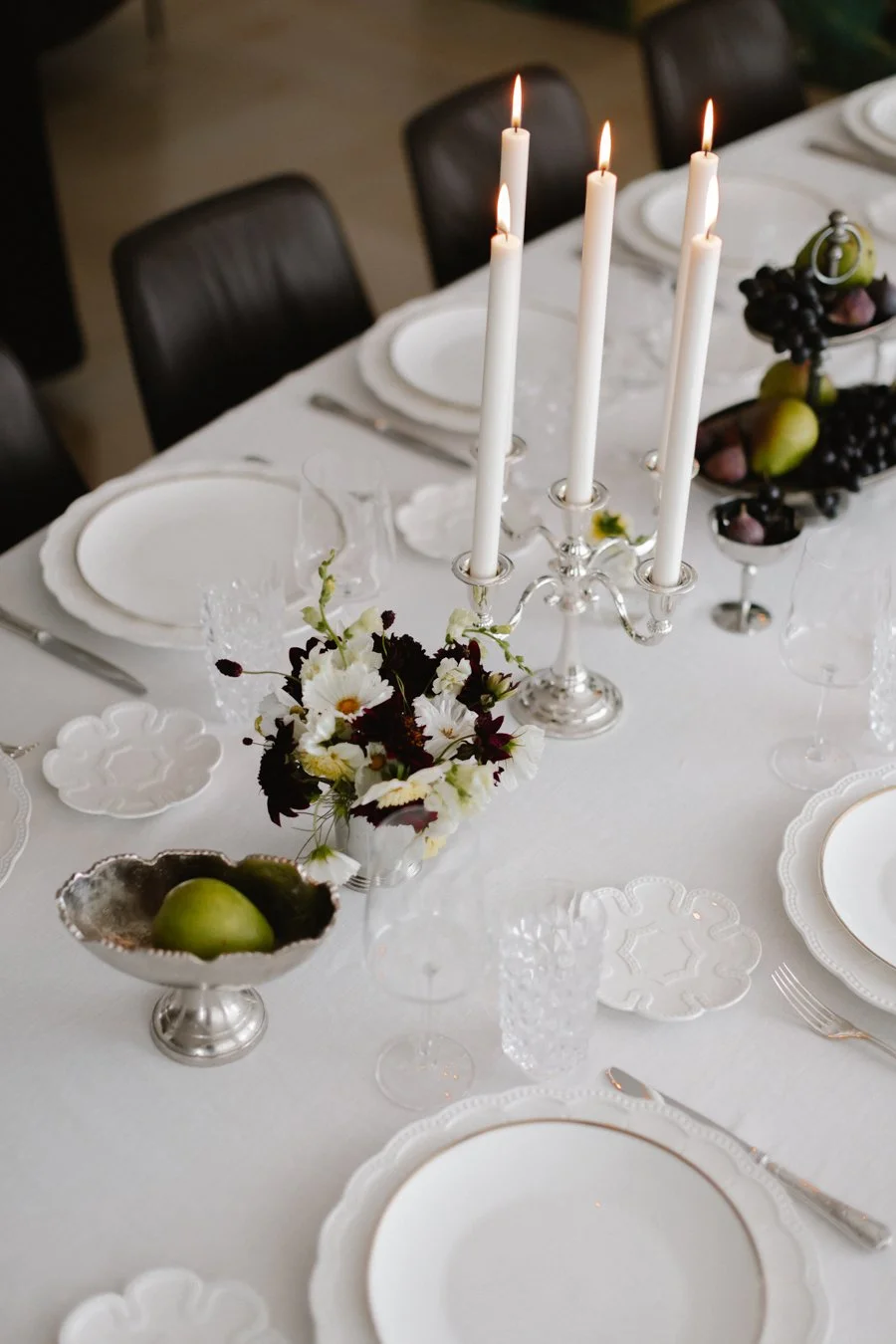 A formal dining table set with white plates, silverware, crystal glasses, a silver candelabra with lit white candles, a small vase with white flowers and dark leaves, a silver bowl with green apples, and a fruit arrangement with grapes and pears.