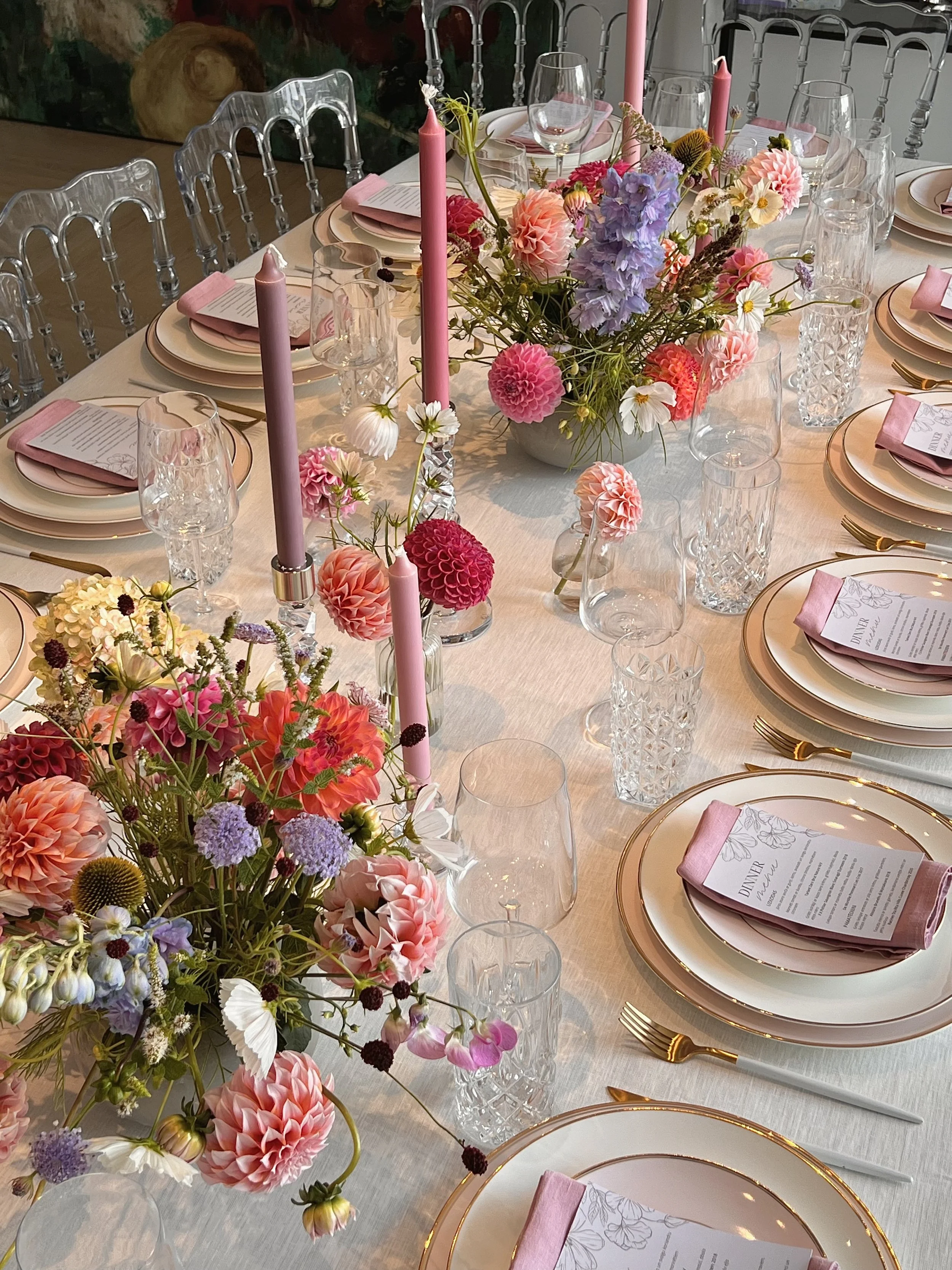 A long dining table set for a formal event with elegant tableware, pink candles, colorful flower arrangements, and pink napkins on white plates with gold rims.