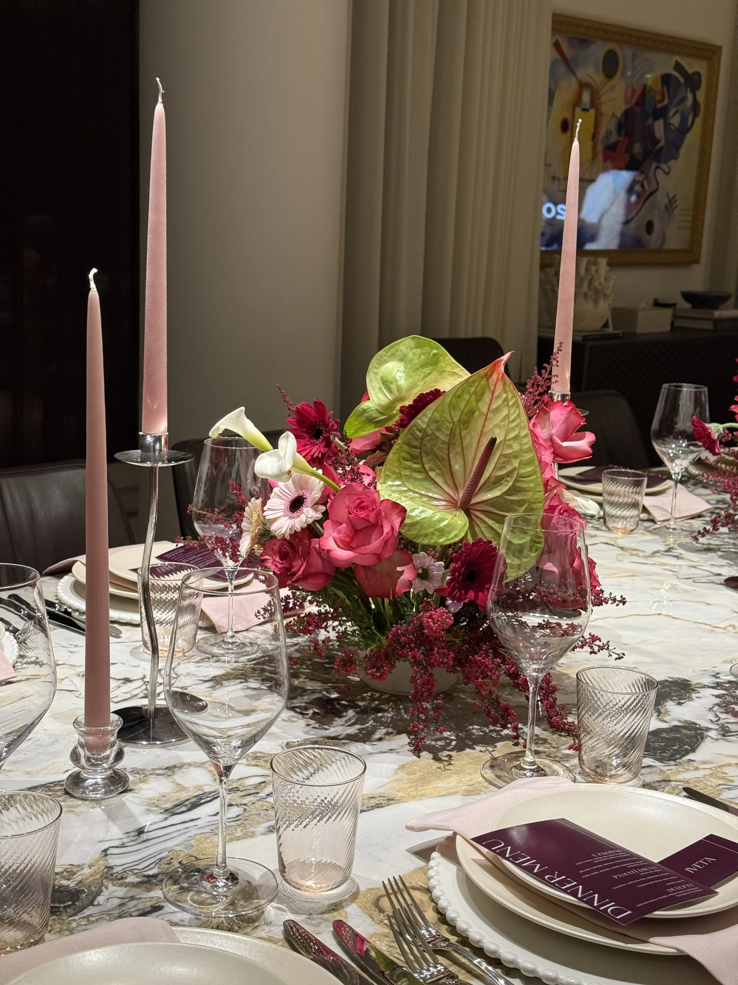 Elegant dining table decorated with a floral centerpiece, pink taper candles, wine glasses, utensils, plates, and menus, set in a dining room with artwork on the wall.
