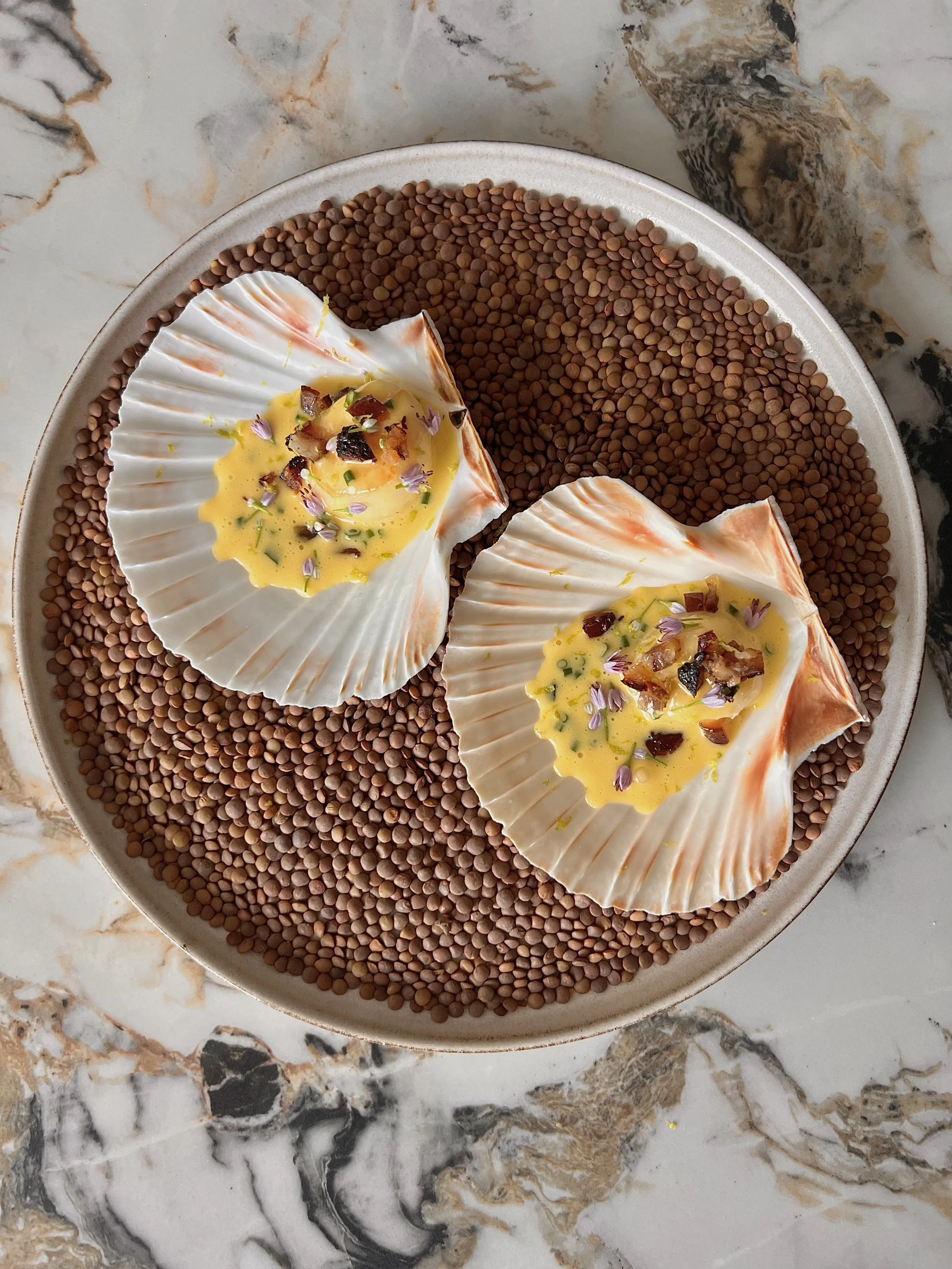 A round plate with two scallop shells filled with creamy Yellow egg custard topped with chopped vegetables and garnished with small purple flowers, placed on a bed of small brown lentils on a marble surface.