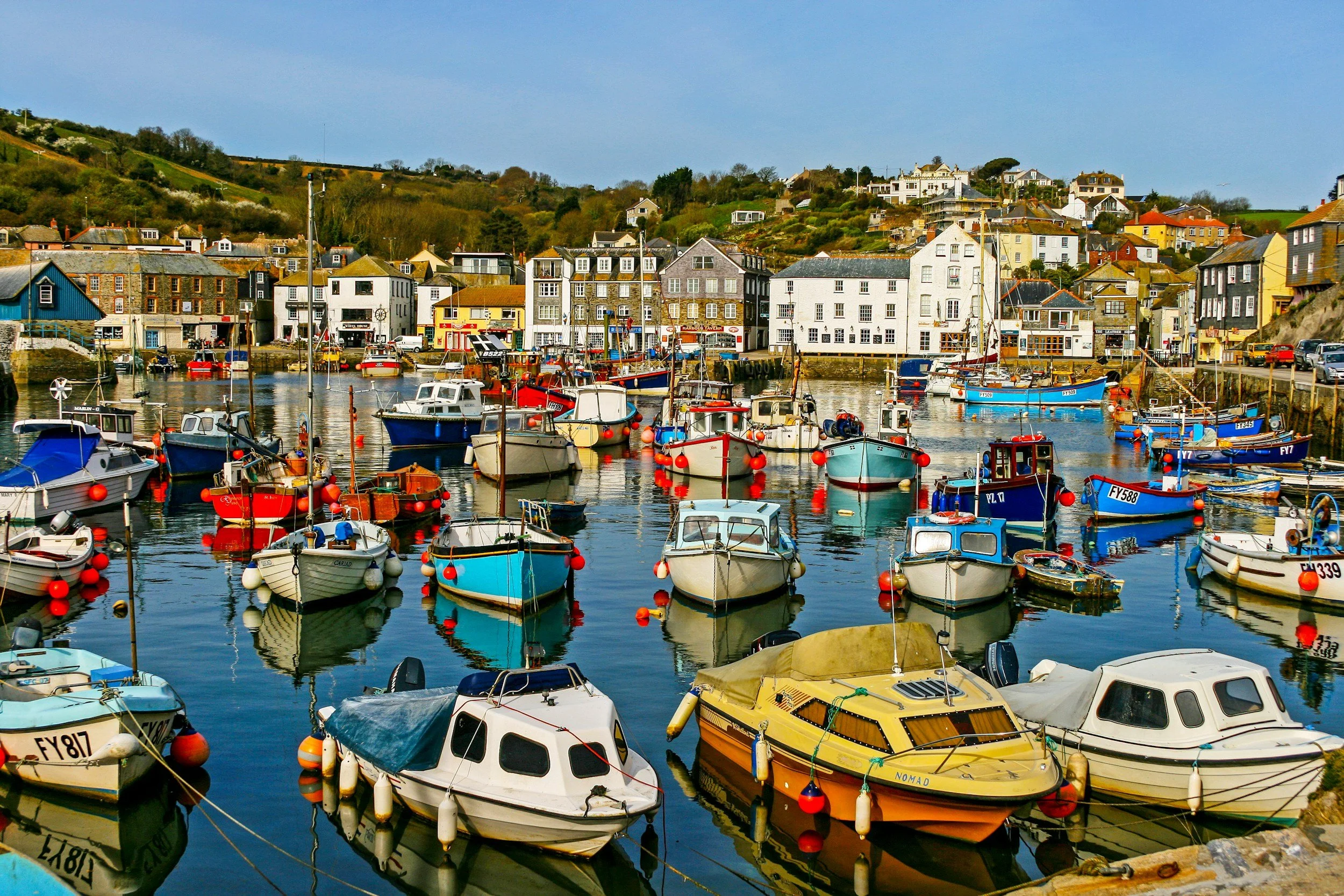 Padstow harbor with various boats docked, surrounded by colorful buildings on the shoreline, and hills with houses in the background.