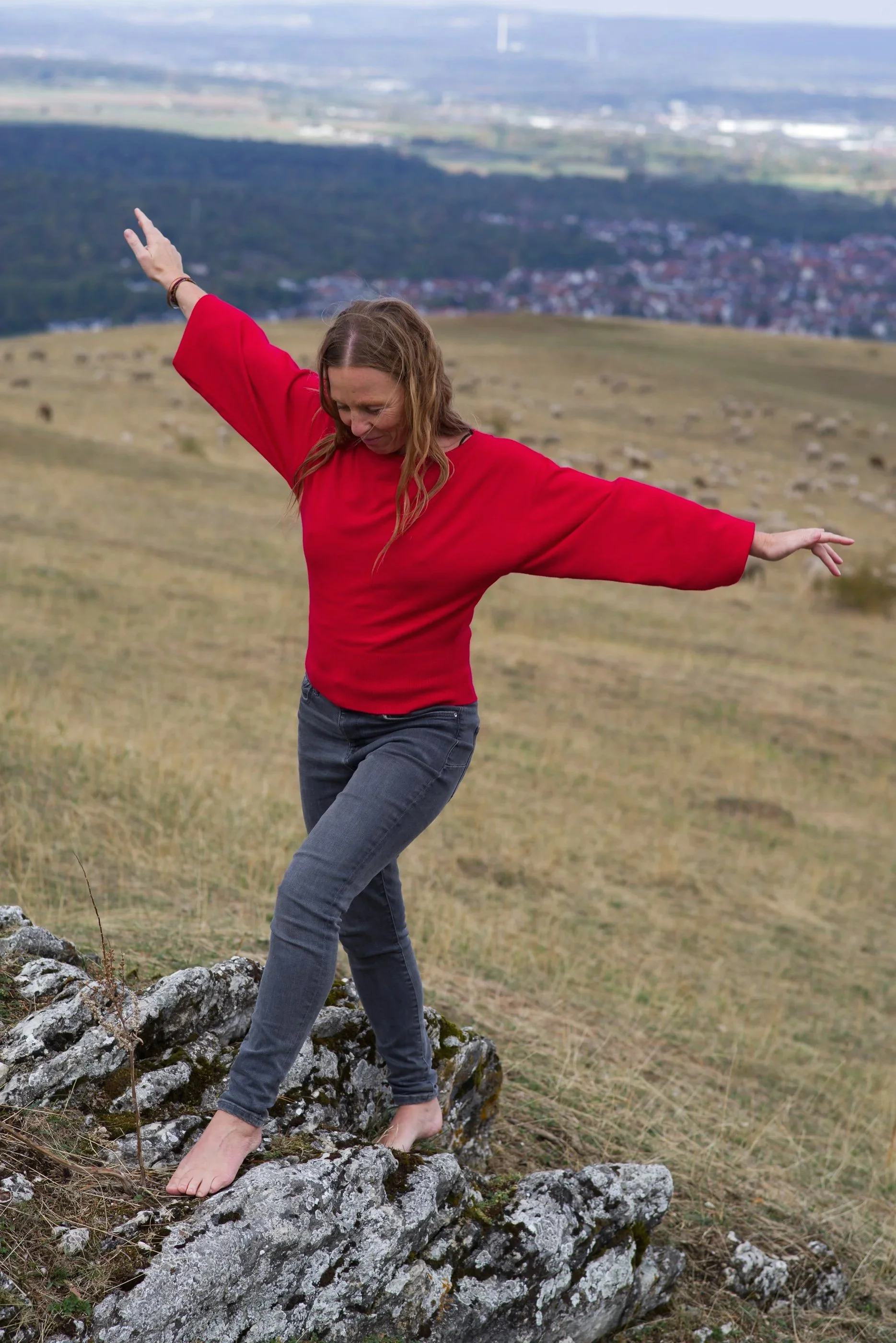 Eine Frau in einem roten Pullover balanciert, sicher und sichtlich zufrieden auf einem Berg. Im HIntergrund kann man verschwommen SChafe und winzige Häuser entdecken.