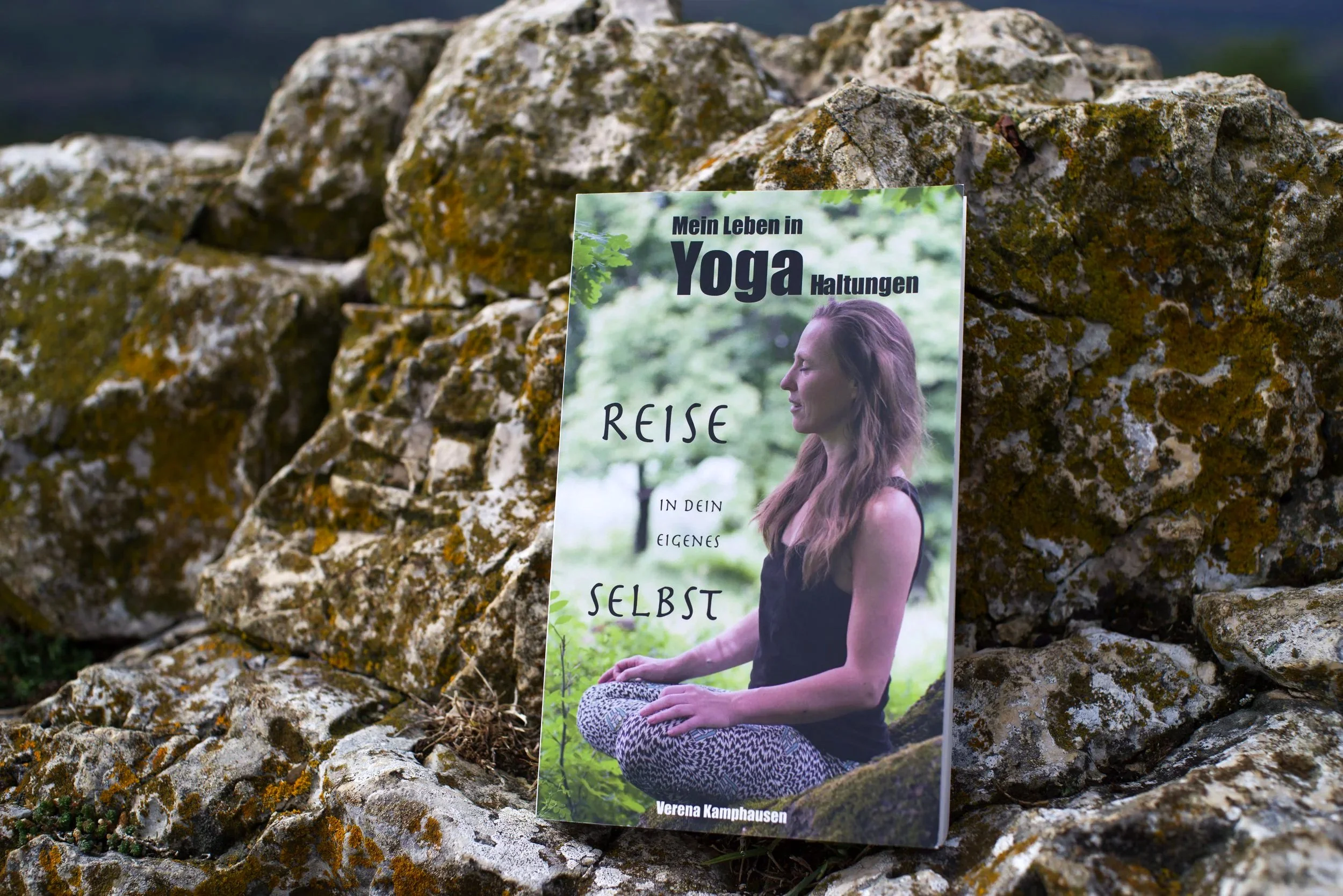 Ein Buch lehnt an einem Fels. Auf dem Cover sieht man eine Frau in Meditationshaltung. Der Titel des Buches lautet: "Mein Leben in Yoga Haltungen. Reise in dein eigenes Selbst."