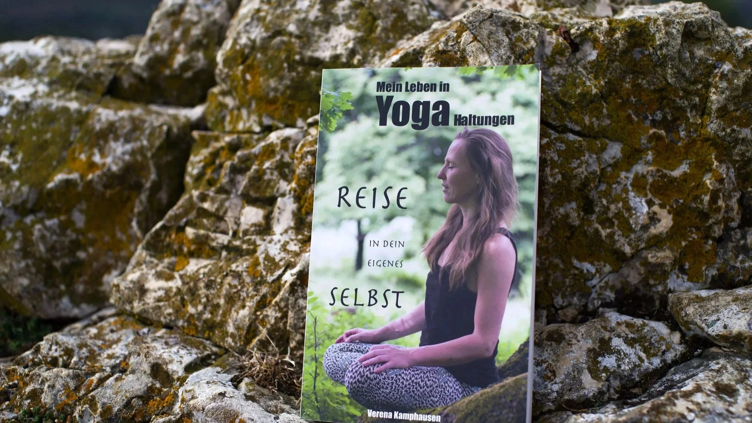 Ein Buch mit dem Titel: "Mein Leben in Yoga Haltungen - Reise in dein eigenes Selbst" lehnt an einem Felsen und wird von der Sonne angeleuchtet