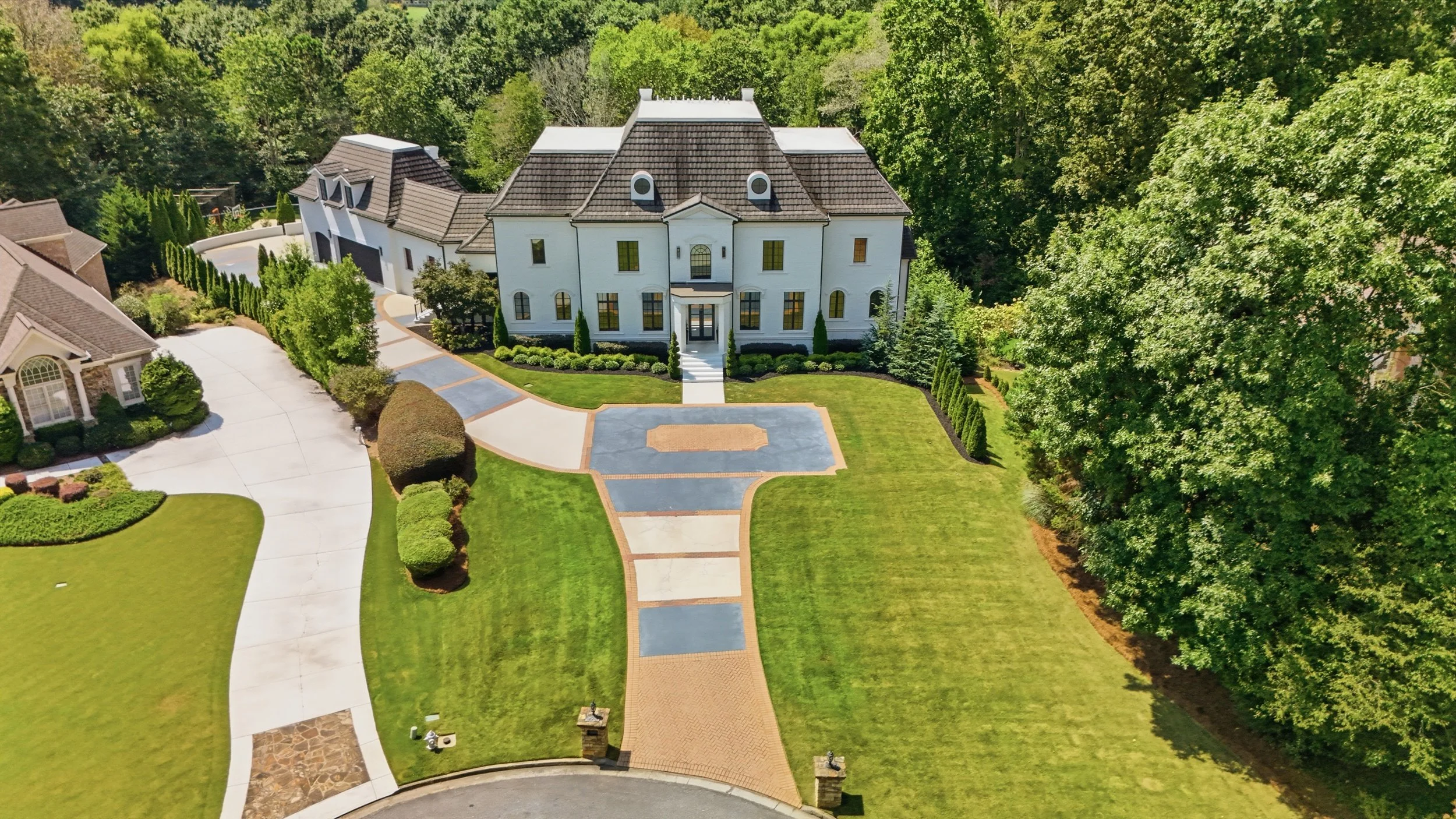 Aerial view of a large, elegant white mansion surrounded by a lush green yard with a curved driveway, well-kept lawn, and decorative shrubs.