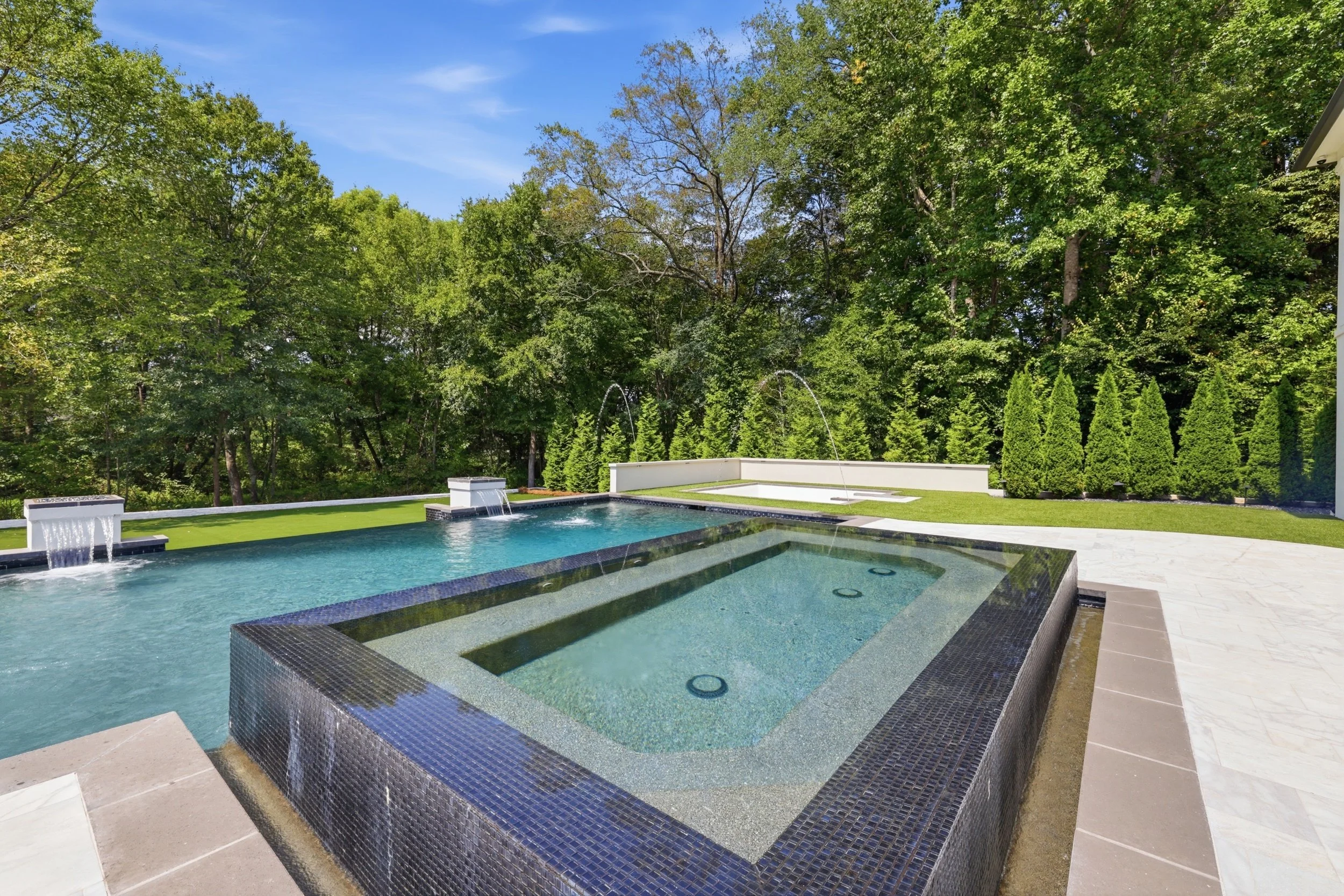A backyard view of a rectangular swimming pool with water features, surrounded by a lush green lawn and tall trees under a blue sky.