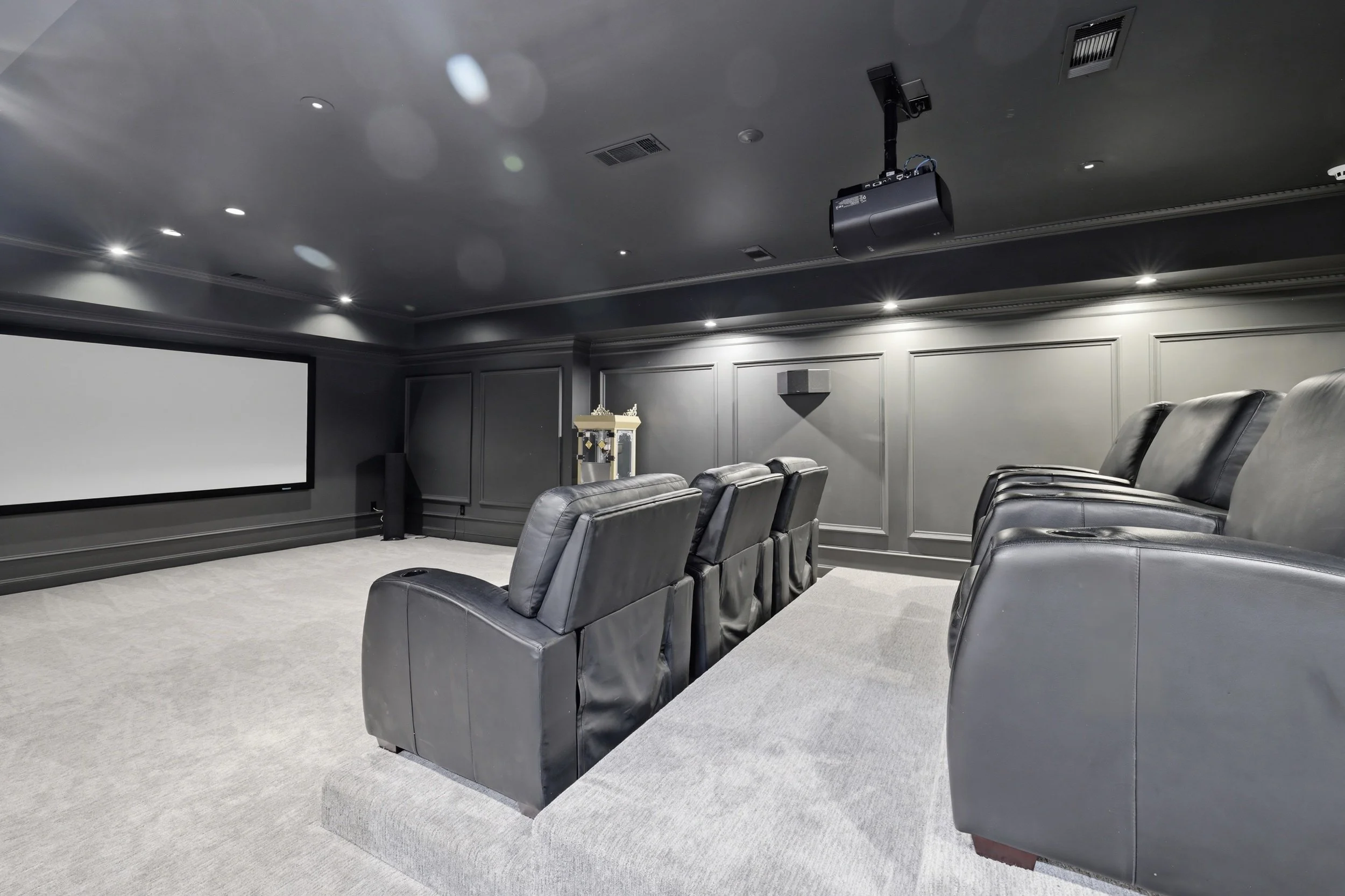 A home theater room with a large white screen, black leather recliner chairs, and a ceiling-mounted projector.