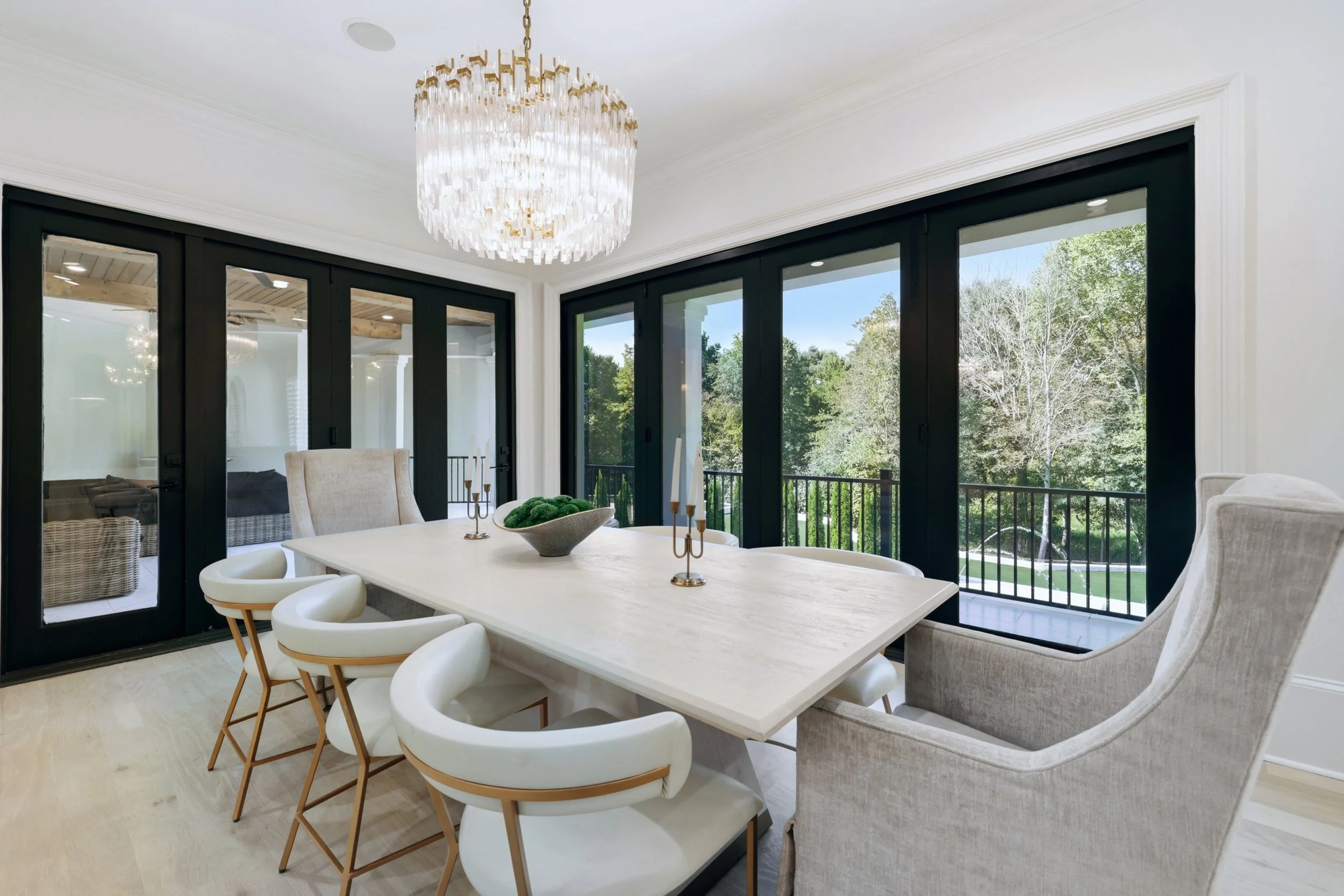 Bright dining room with a white table, six modern white chairs, and an elegant chandelier above. Large black-framed sliding glass doors reveal an outdoor patio and greenery.