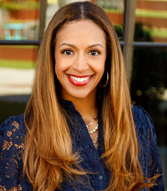 A woman with long, light brown hair and brown eyes, wearing a navy blue lace blouse, gold hoop earrings, and layered necklaces, smiling in front of a window.