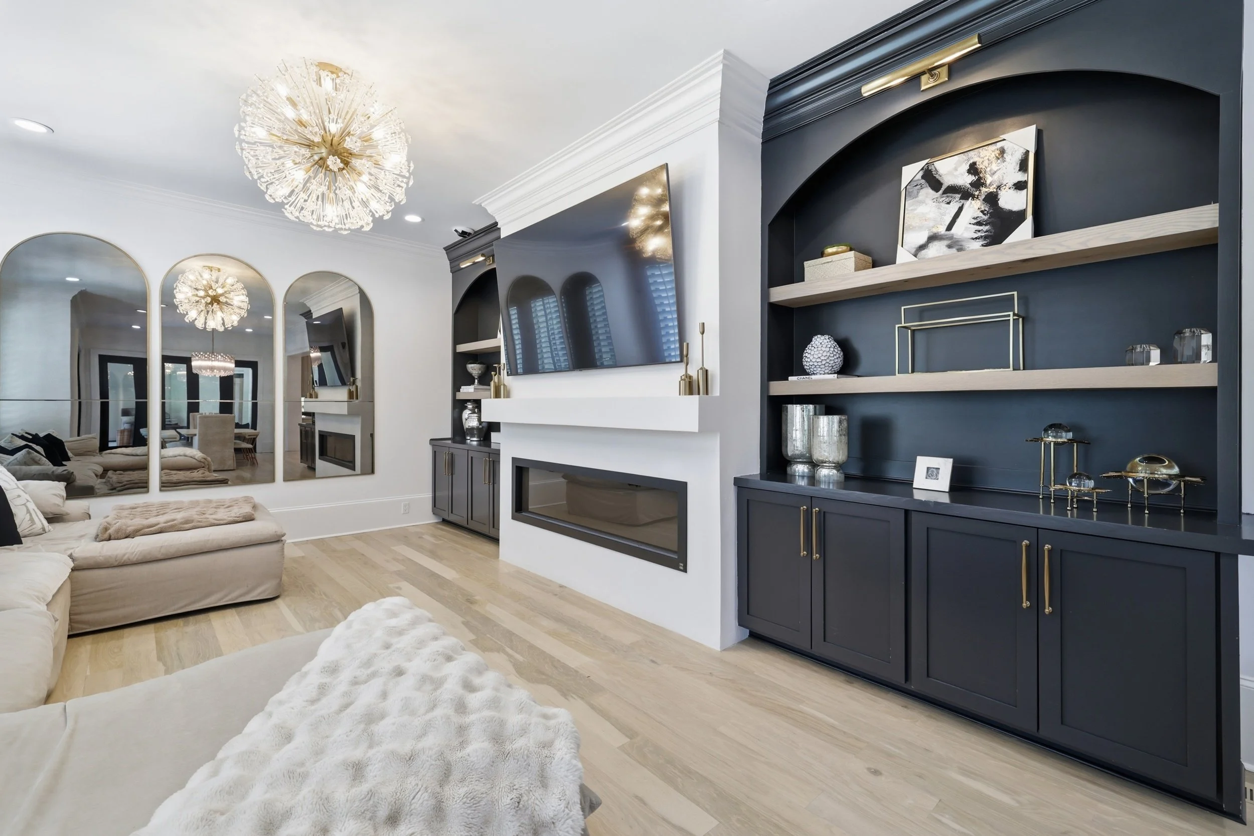 A modern living room featuring a beige sectional sofa, a white textured rug, a black accent wall with built-in shelves, a large flat-screen TV, and decorative items. There is a chandelier with multiple light fixtures on the ceiling and three large ar