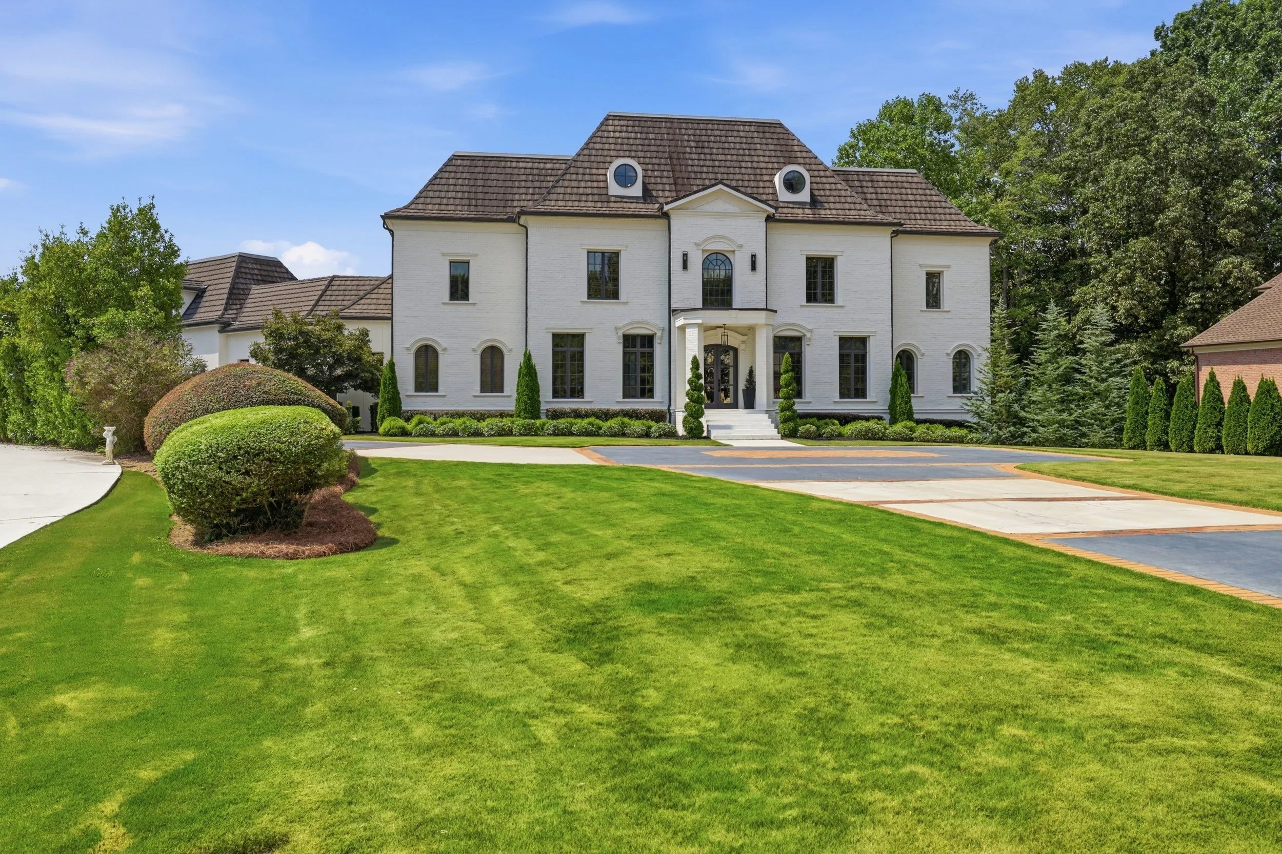 A large white house with a brown shingle roof, multiple windows, and a small front porch. The house is surrounded by well-maintained green grass, bushes, and trees, under a clear blue sky.