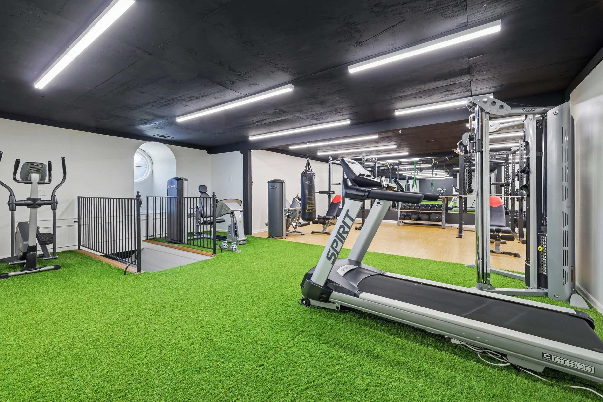 Inside a modern gym with various fitness equipment, including a treadmill, boxing punching bag, elliptical machine, and weightlifting area, with a green artificial turf section and a wood floor area, illuminated by bright ceiling lights.