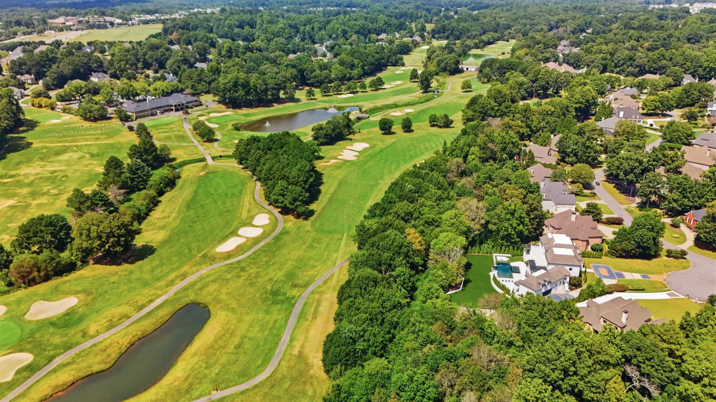Aerial view of a golf course with green fairways, sand traps, water hazards, surrounded by residential houses and trees.