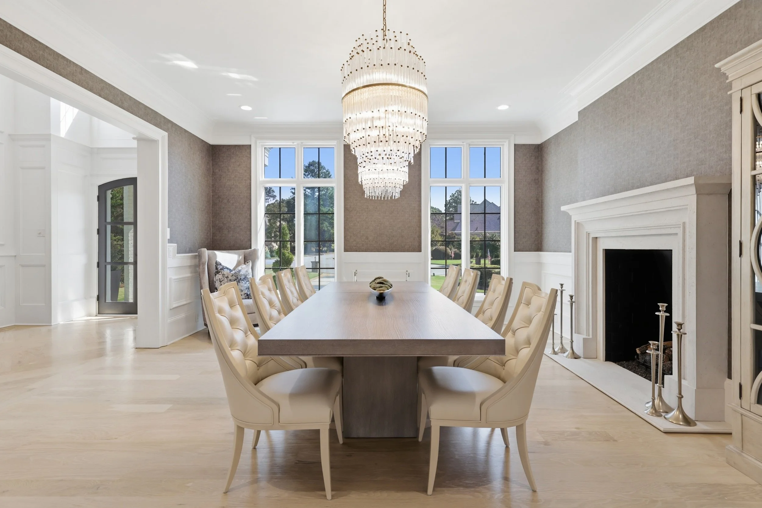 Elegant dining room with a long wooden table surrounded by cream-colored tufted chairs, a modern chandelier hanging from the ceiling, large windows providing natural light, and a fireplace with candles on the hearth.