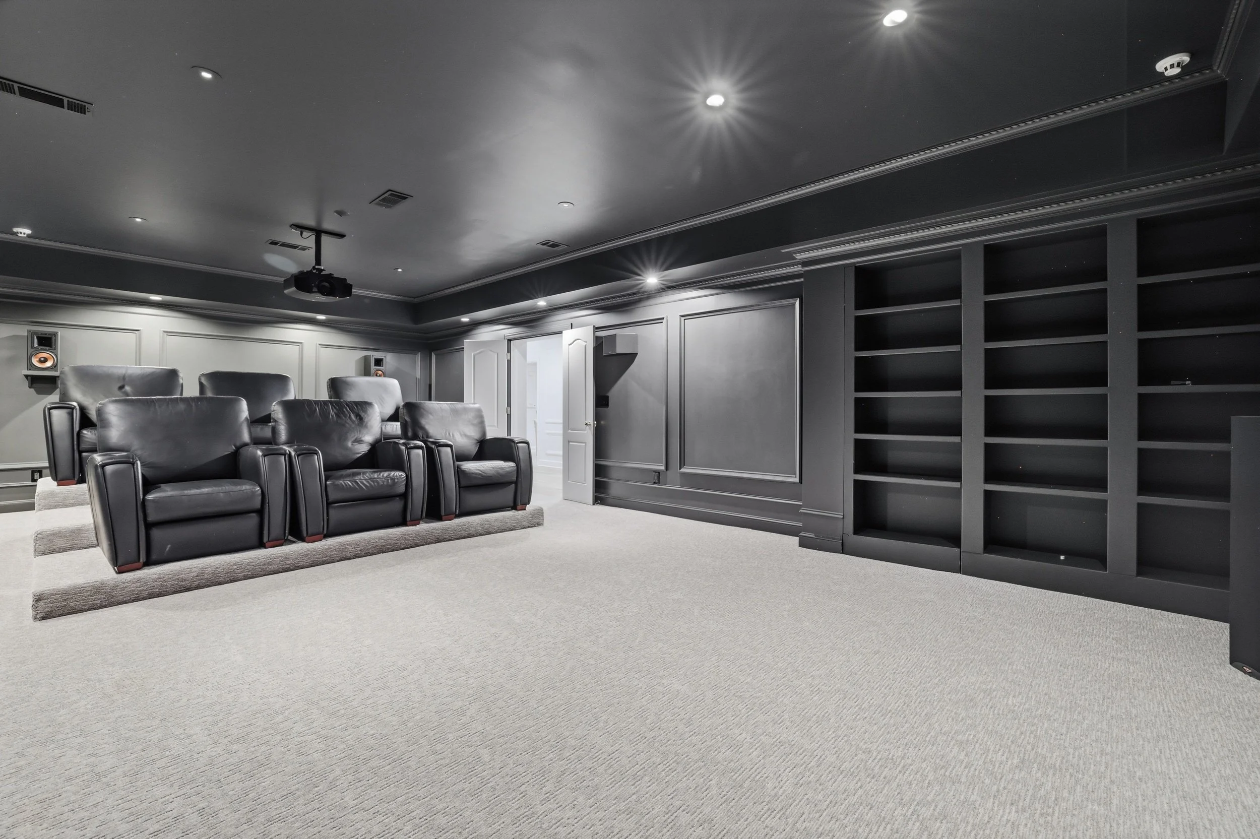 Dark gray home theater room with black leather seats, a wall of empty black shelves, and a ceiling mounted projector.
