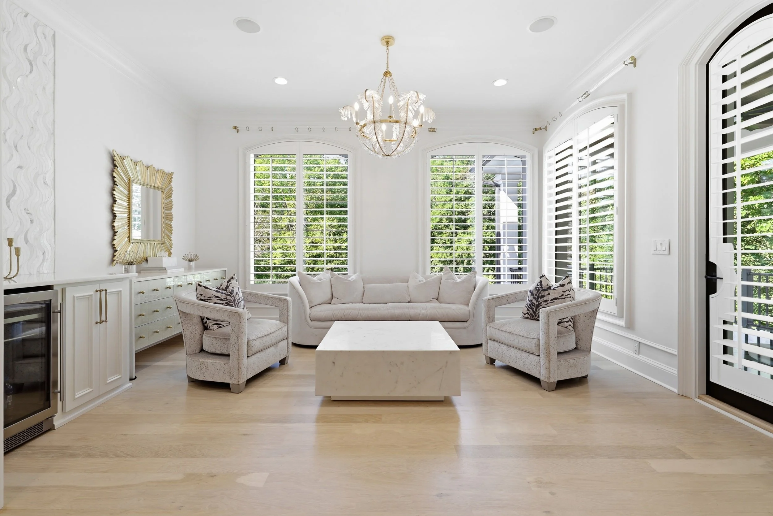 Bright living room with white walls, large windows with white shutters, and a beige sofa with pillows. Decor includes a gold-framed mirror, gold accents, and a chandelier with feather-like decorations.