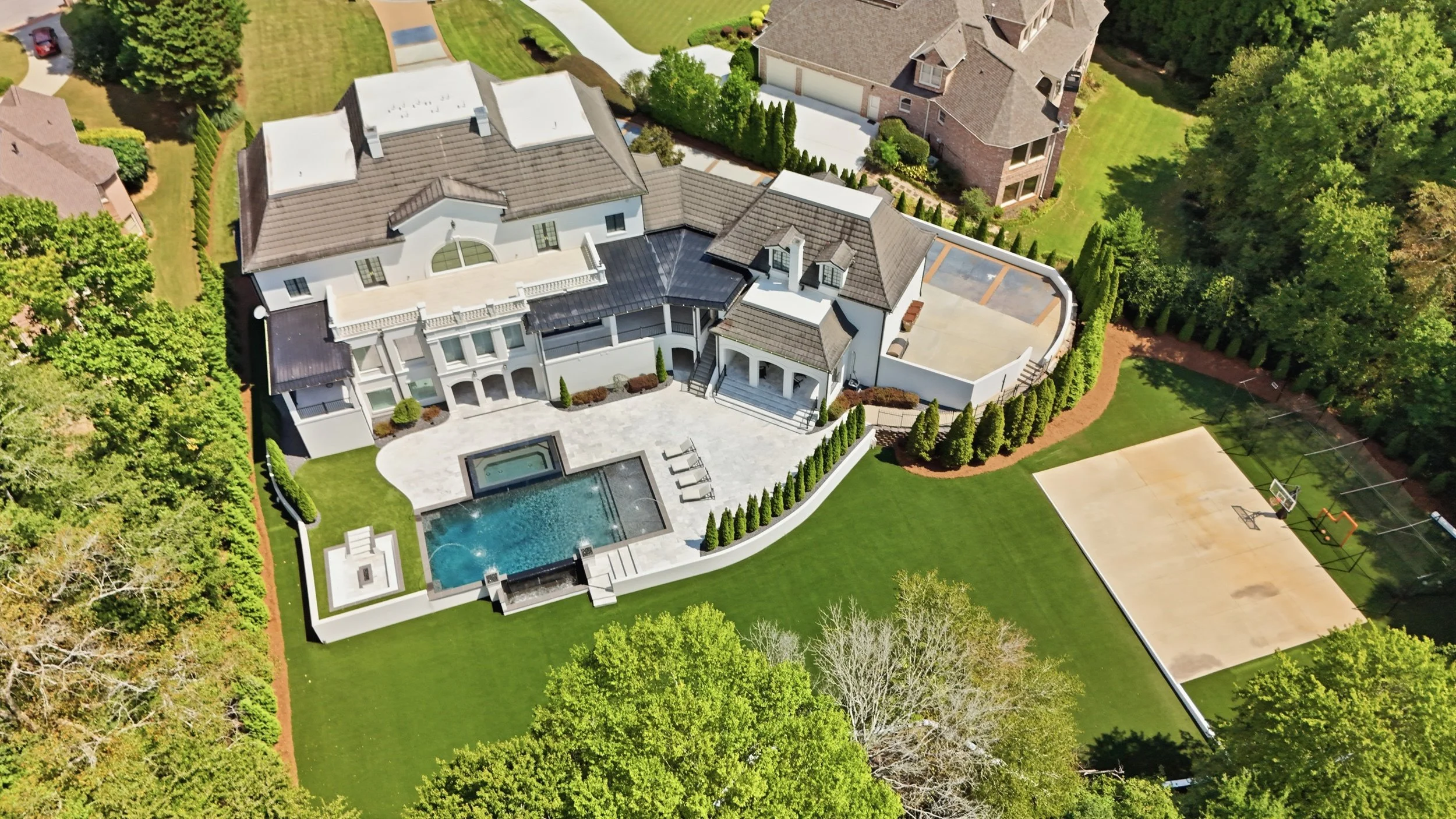 Aerial view of a large, modern house with a backyard, swimming pool, basketball court, and surrounding greenery.