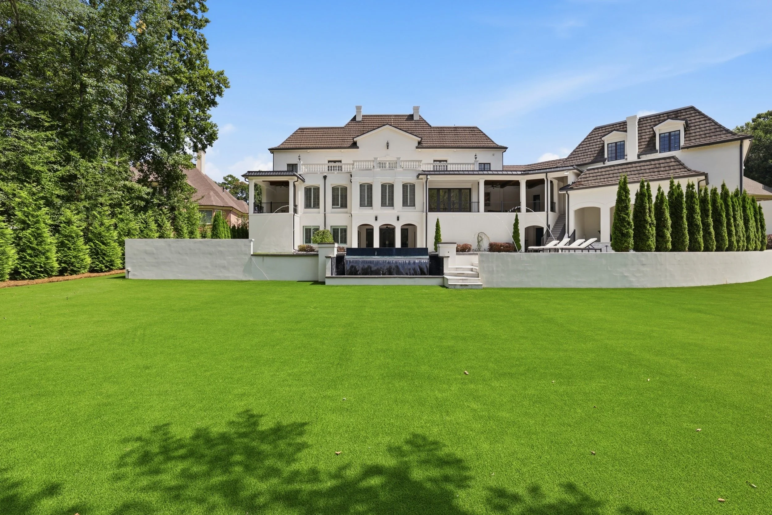 Large white mansion with multiple levels, balconies, and white shutters, surrounded by a lush green lawn and tall trees, under a clear blue sky.