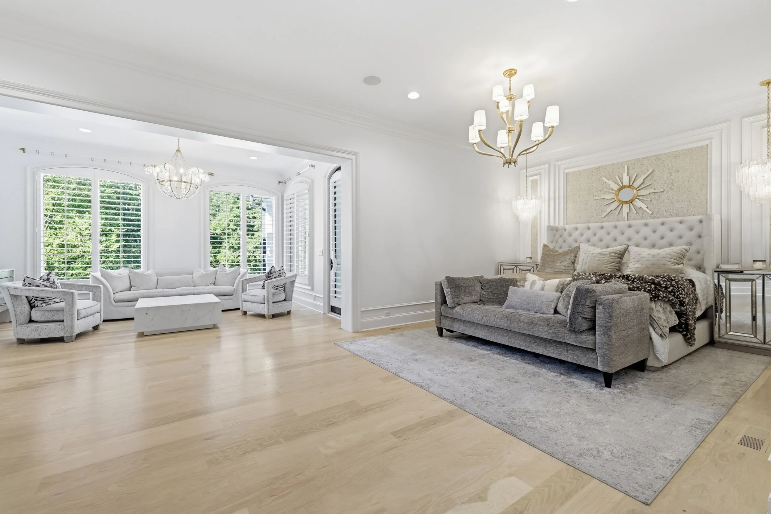 A bright, modern living room with white walls, light wood flooring, and white window shutters. There are two chandeliers and a large, plush white sofa with patterned pillows on a gray rug, along with a smaller gray sofa and a side table.