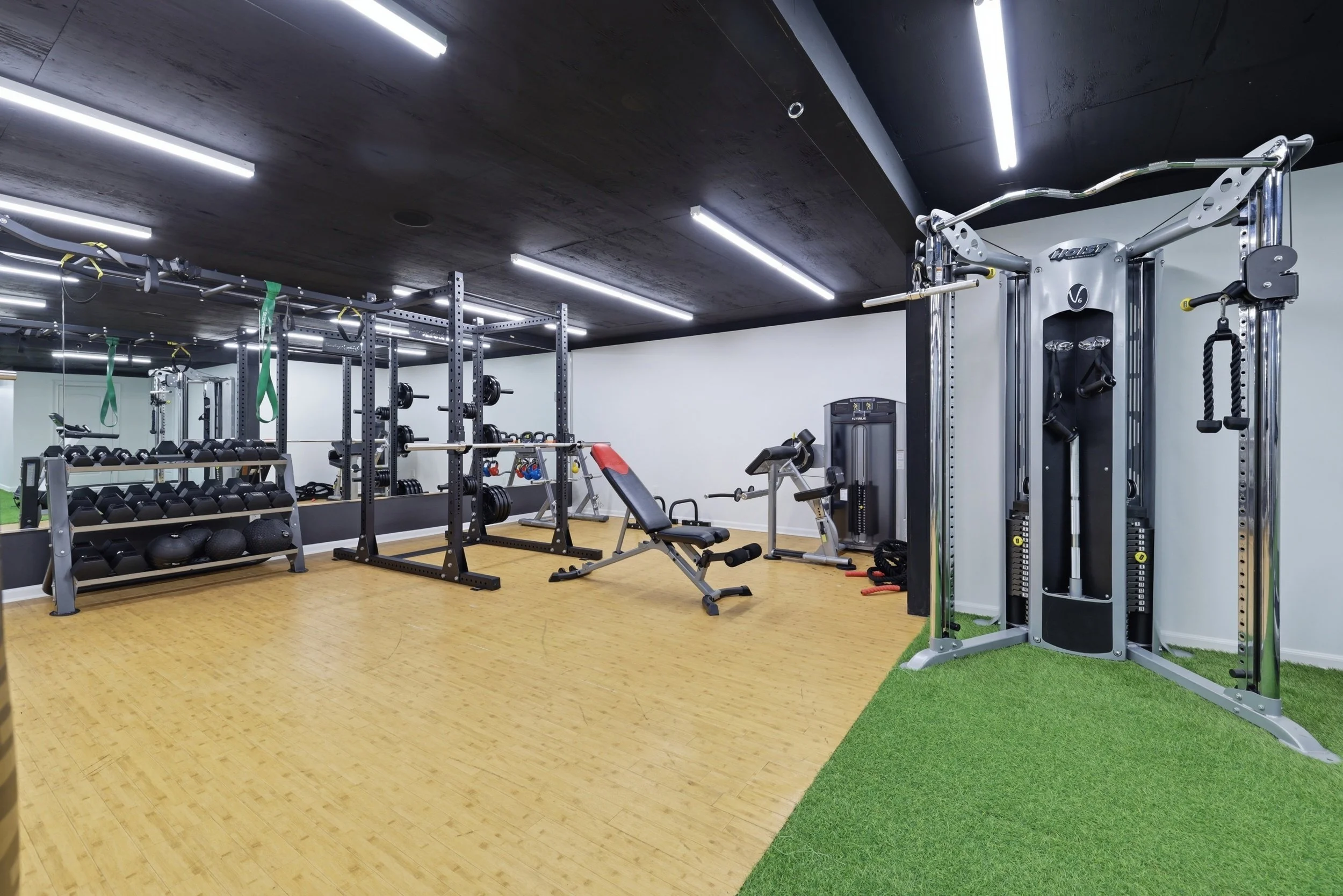A modern gym with various workout equipment including dumbbells, barbells, benches, resistance bands, and a power rack. The flooring is wooden, and the ceiling has black panels with LED strip lighting.