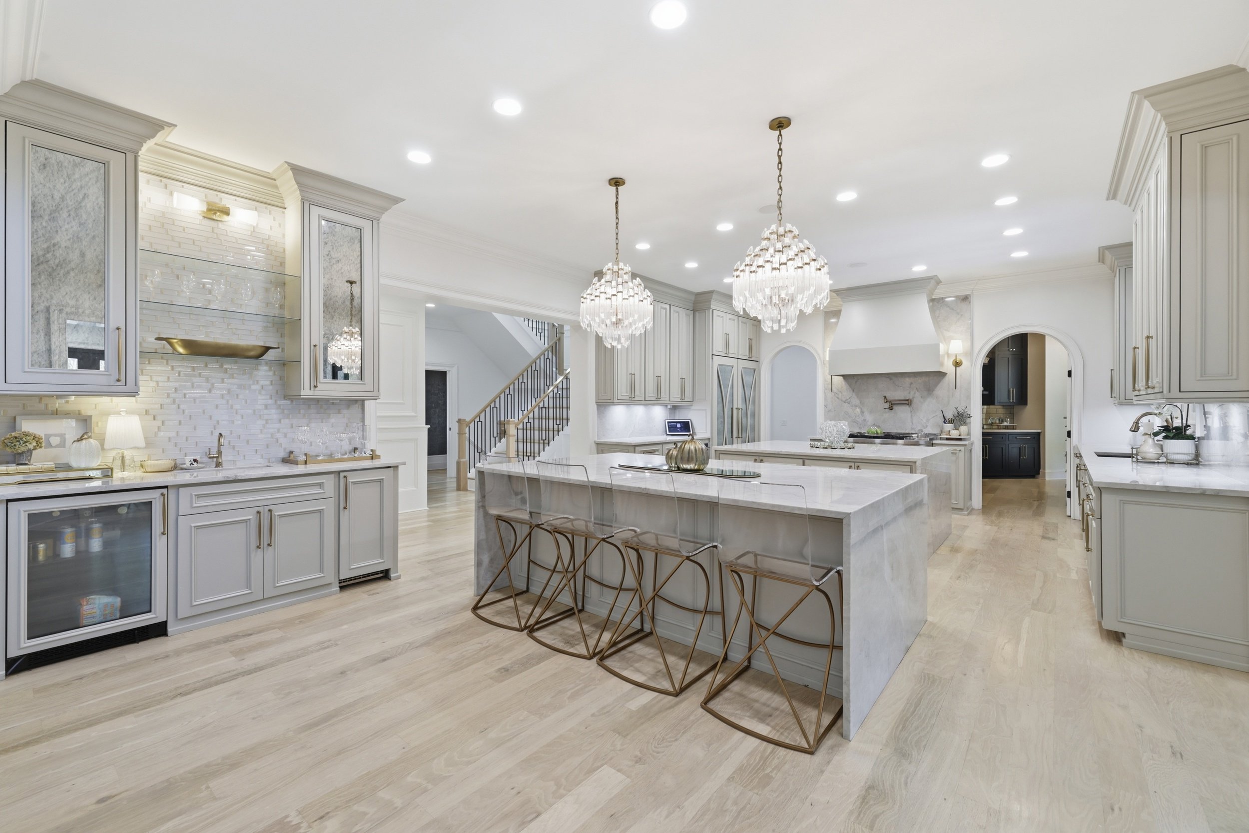 A spacious, modern white kitchen with a large island, elegant hanging chandeliers, light wood flooring, and detailed cabinetry. The space includes a stove, sink, and decorative accents like plants and a golden bowl.