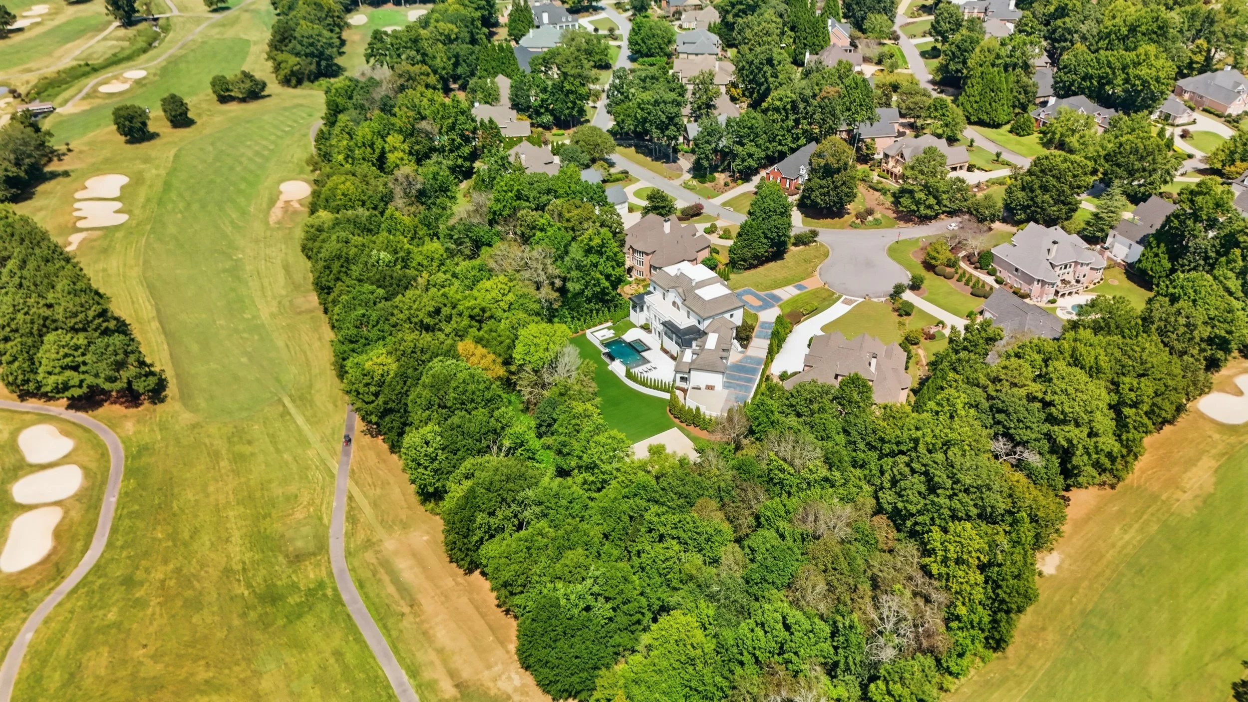 Aerial view of a neighborhood with large houses, green lawns, and many trees, adjacent to a golf course with sand traps and walking paths.
