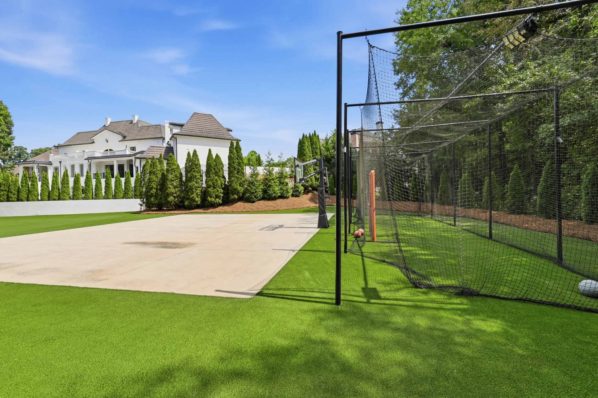 A backyard with a basketball court, a house with white walls and a brown roof, surrounded by green trees and bushes, with a blue sky overhead.