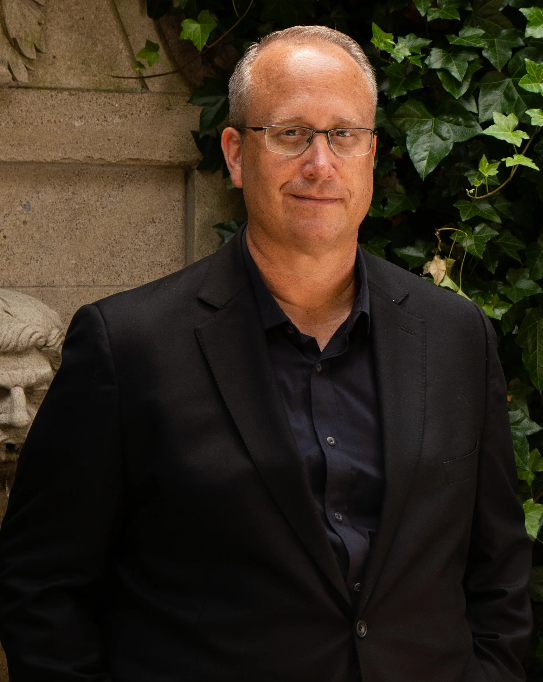 A man with short gray hair wearing glasses, a dark shirt, and a black blazer stands outdoors near a stone wall and green ivy.