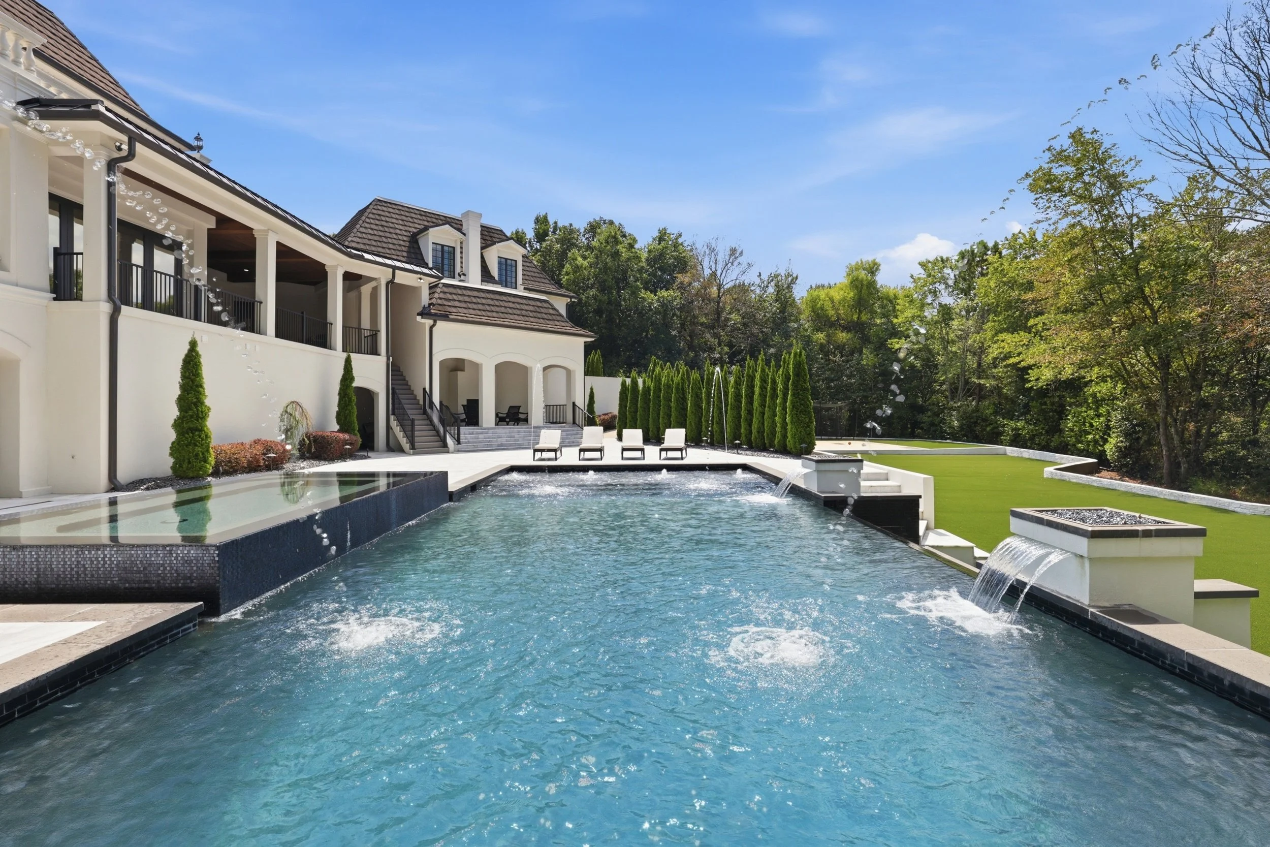 Modern house with a large outdoor swimming pool, surrounded by greenery and lounge chairs, under a blue sky.