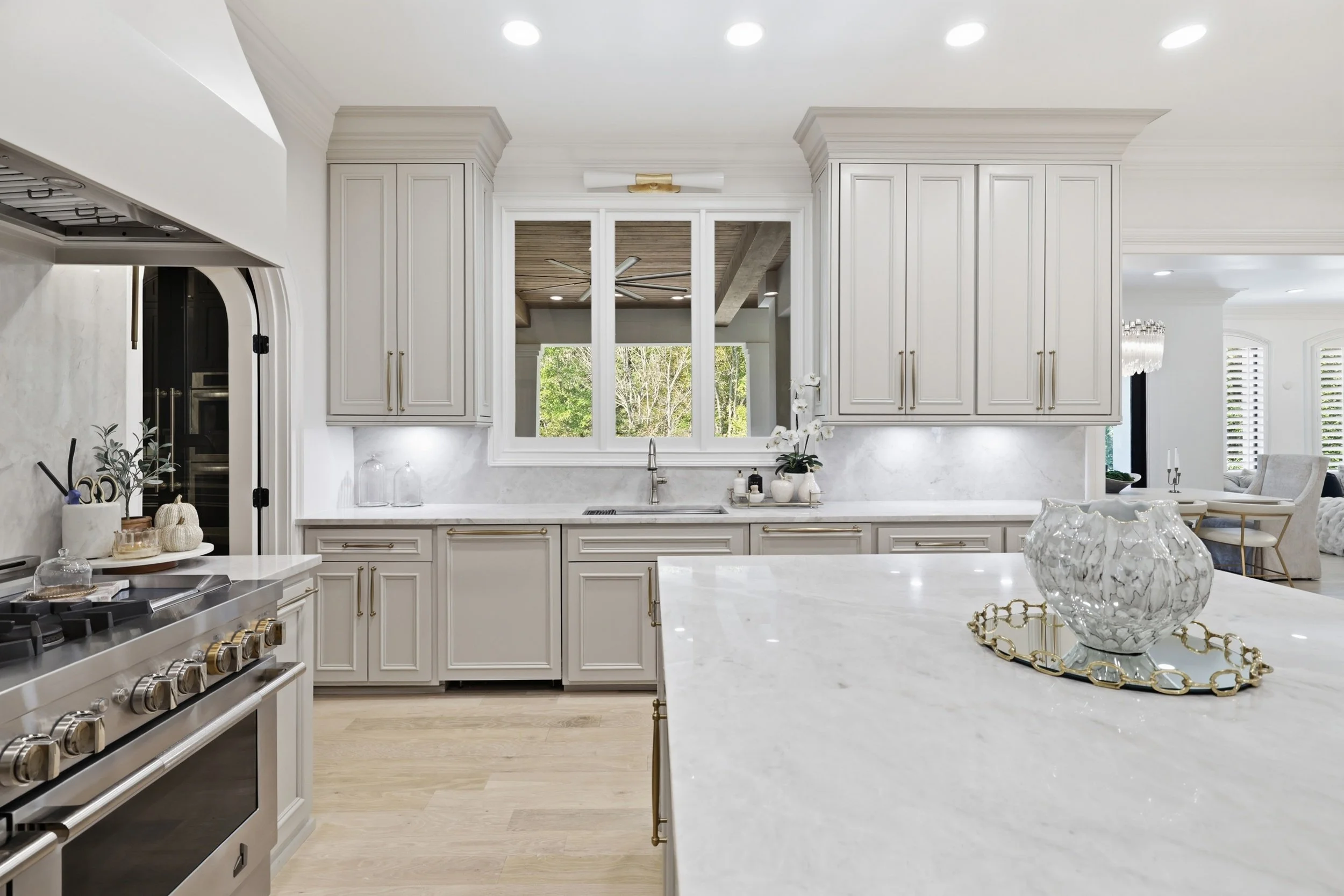 Modern white kitchen with marble countertops, cabinets, and a window overlooking greenery, with stainless steel stove, decorative items, and a view into a living area.