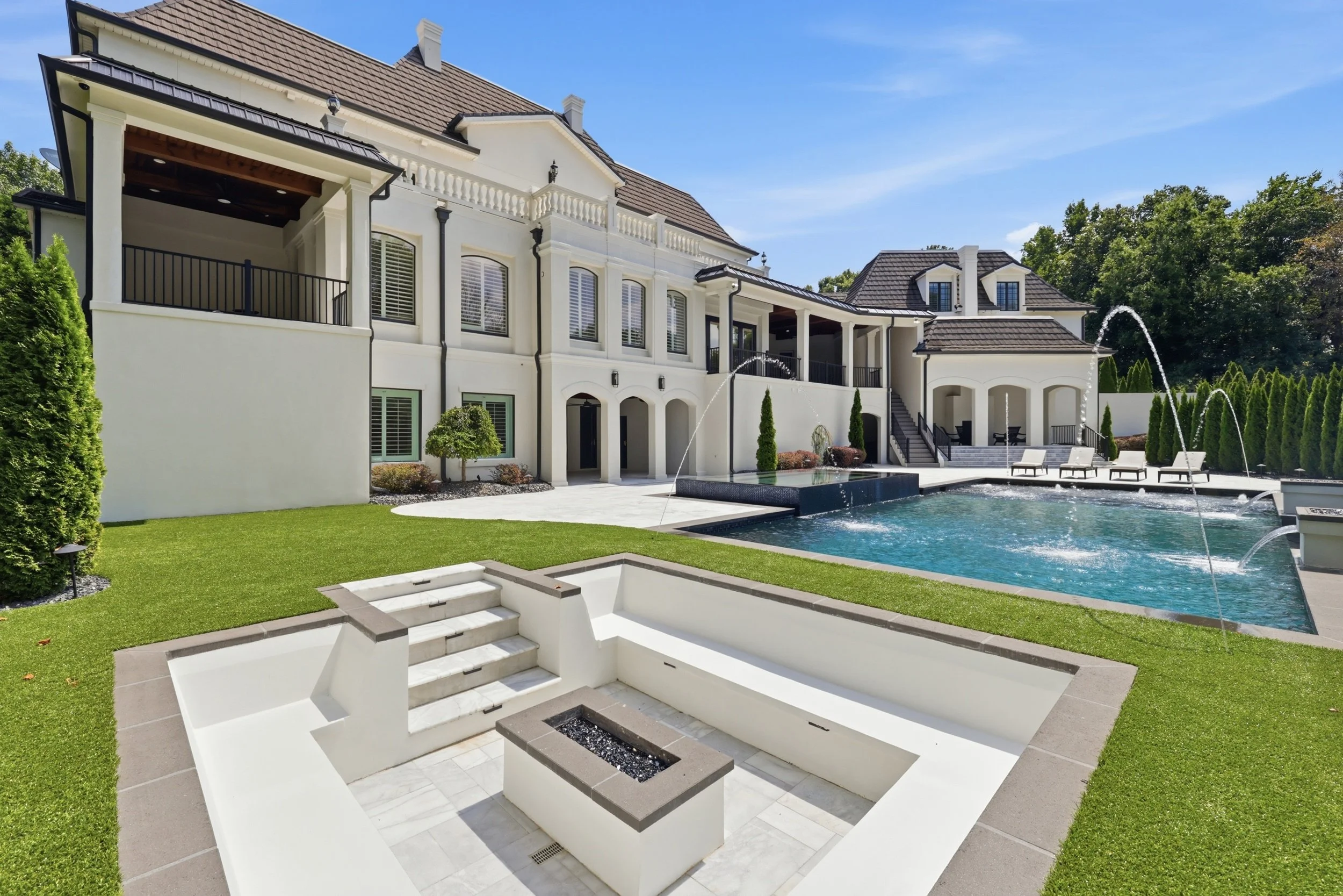 Large, luxurious white house with multiple levels, dark roof, balconies, and large windows, surrounding a backyard with a swimming pool, green lawn, and small trees, under a clear blue sky.