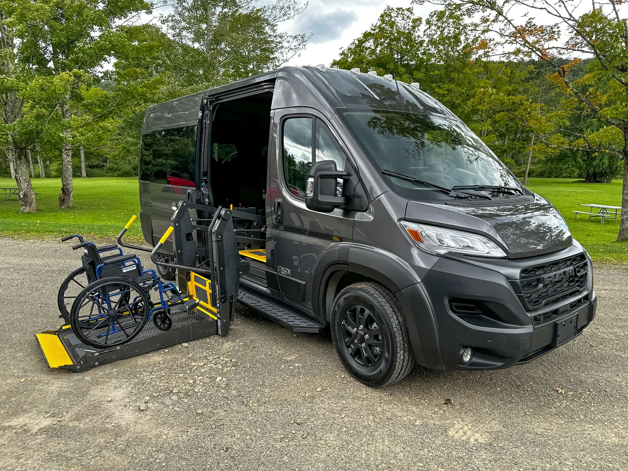 A black van with a wheelchair lift extended at the rear, parked on a grassy area with trees in the background.