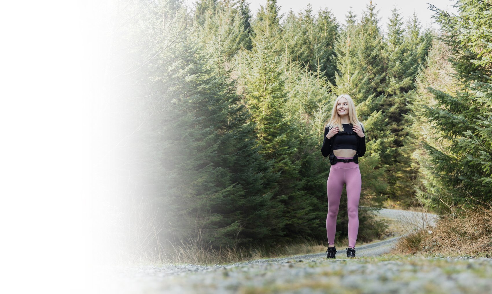A young woman with long blonde hair wearing a black crop top and pink leggings, standing on a trail in a forest of evergreen trees, smiling and looking off into the distance.