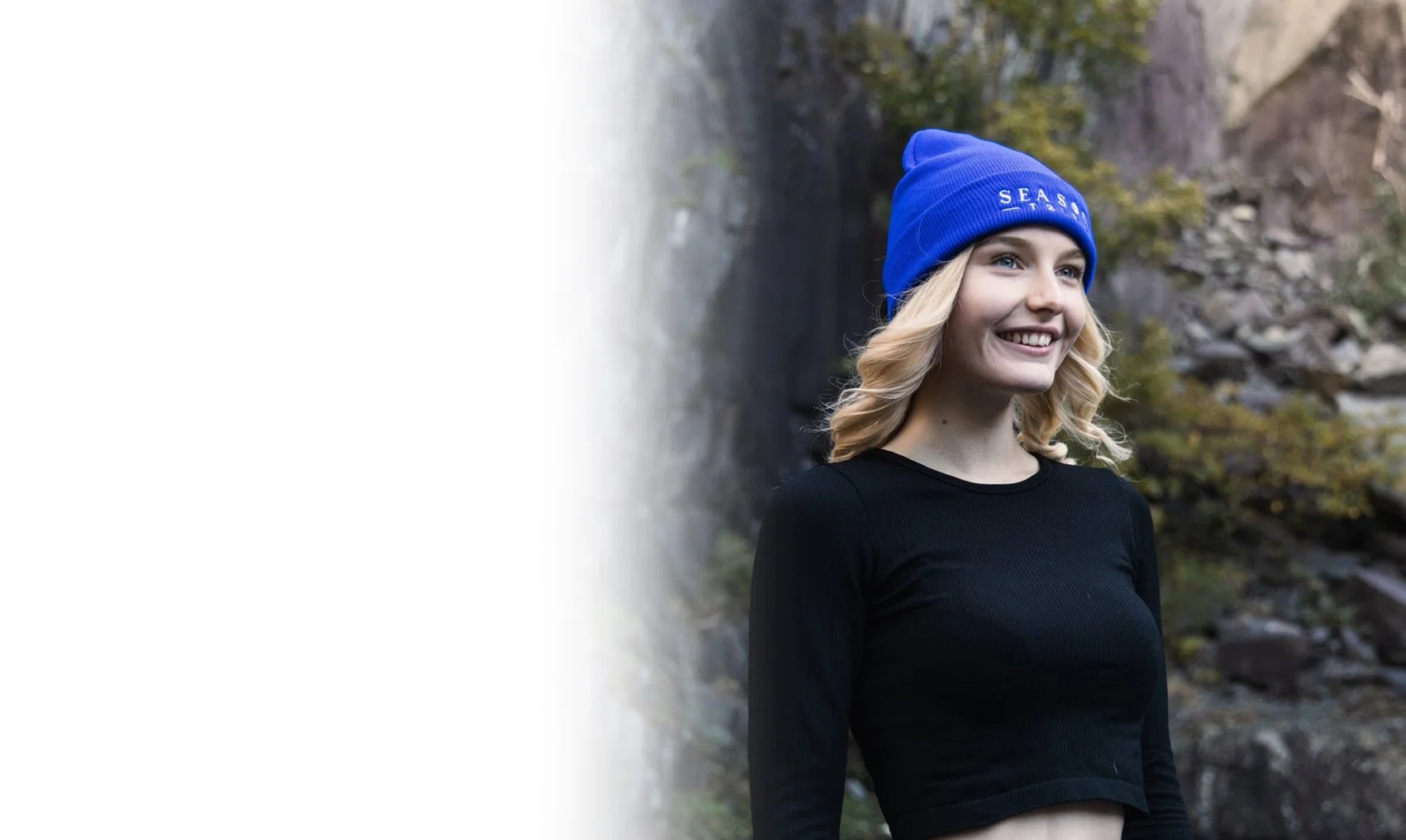 A young woman with blonde hair wearing a blue beanie and a black long-sleeve shirt, smiling outdoors near a waterfall and rocks.