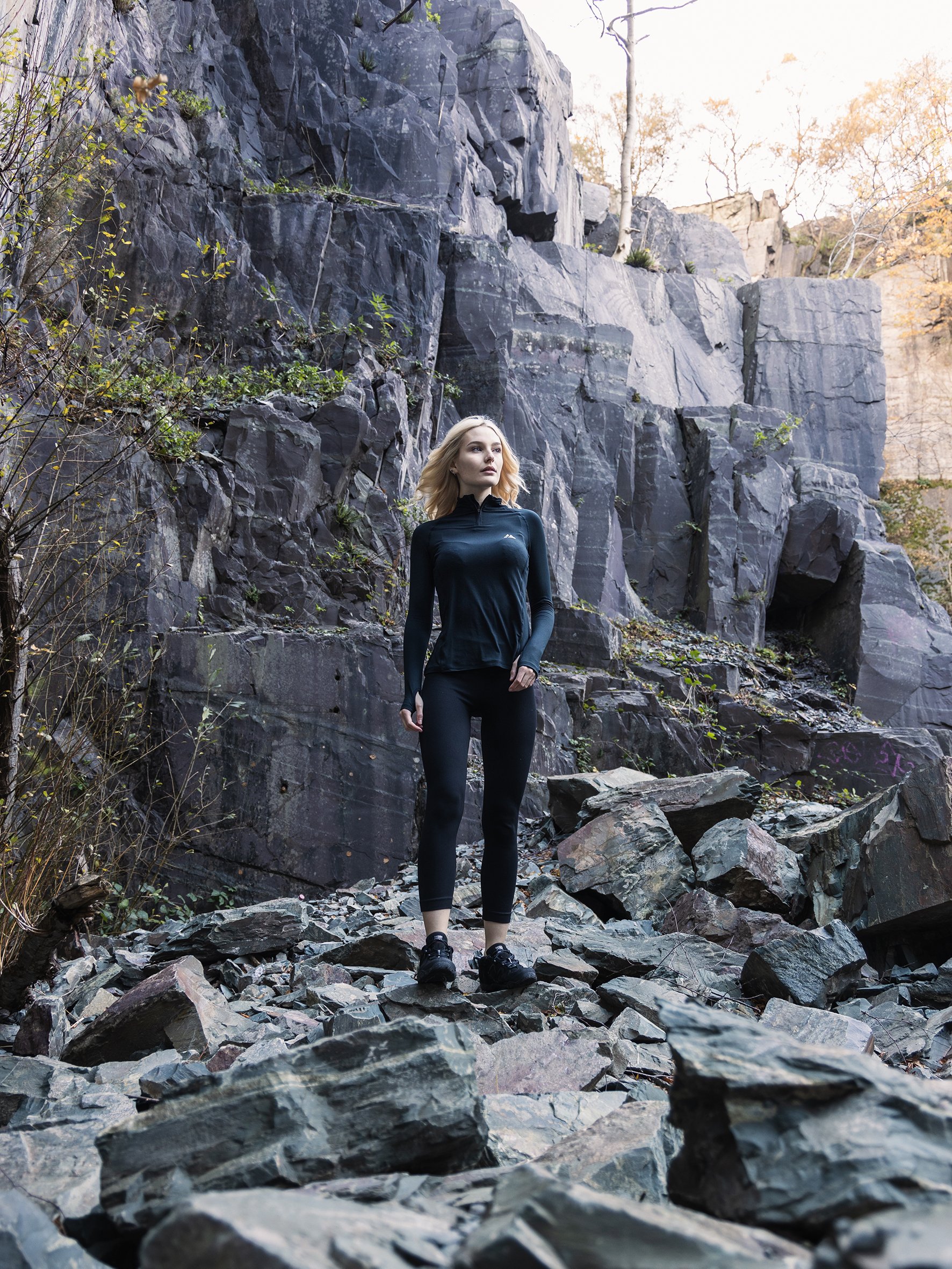 A woman dressed in athletic wear, including black pants and a black long-sleeve shirt, walking on a rocky trail at the base of a steep rocky cliff with sparse vegetation and trees in the background.