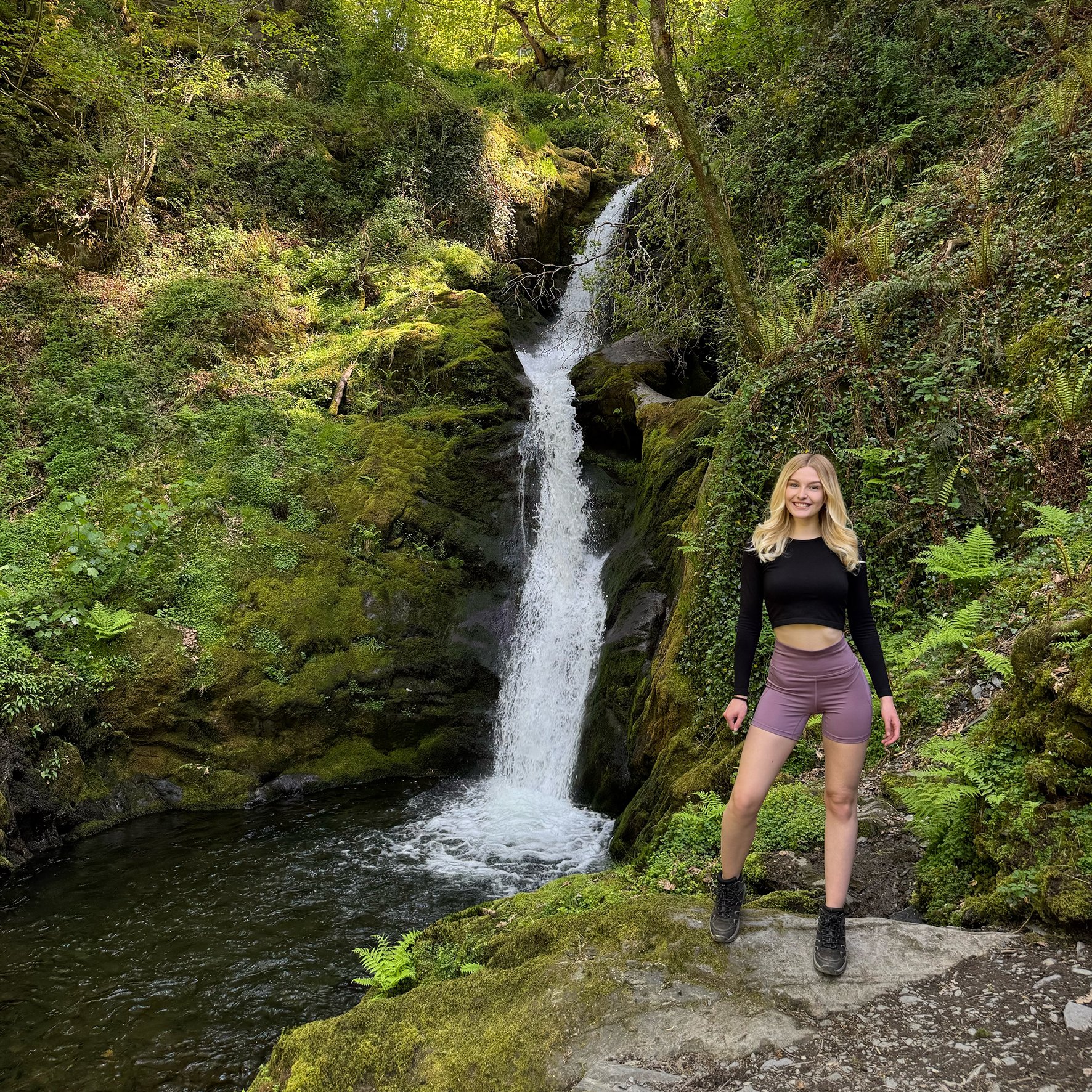 A young woman with long blonde hair wearing a black long sleeve crop top, light purple shorts, and hiking boots standing at the edge of a small stream in a lush forest with a waterfall cascading down moss-covered rocks behind her.
