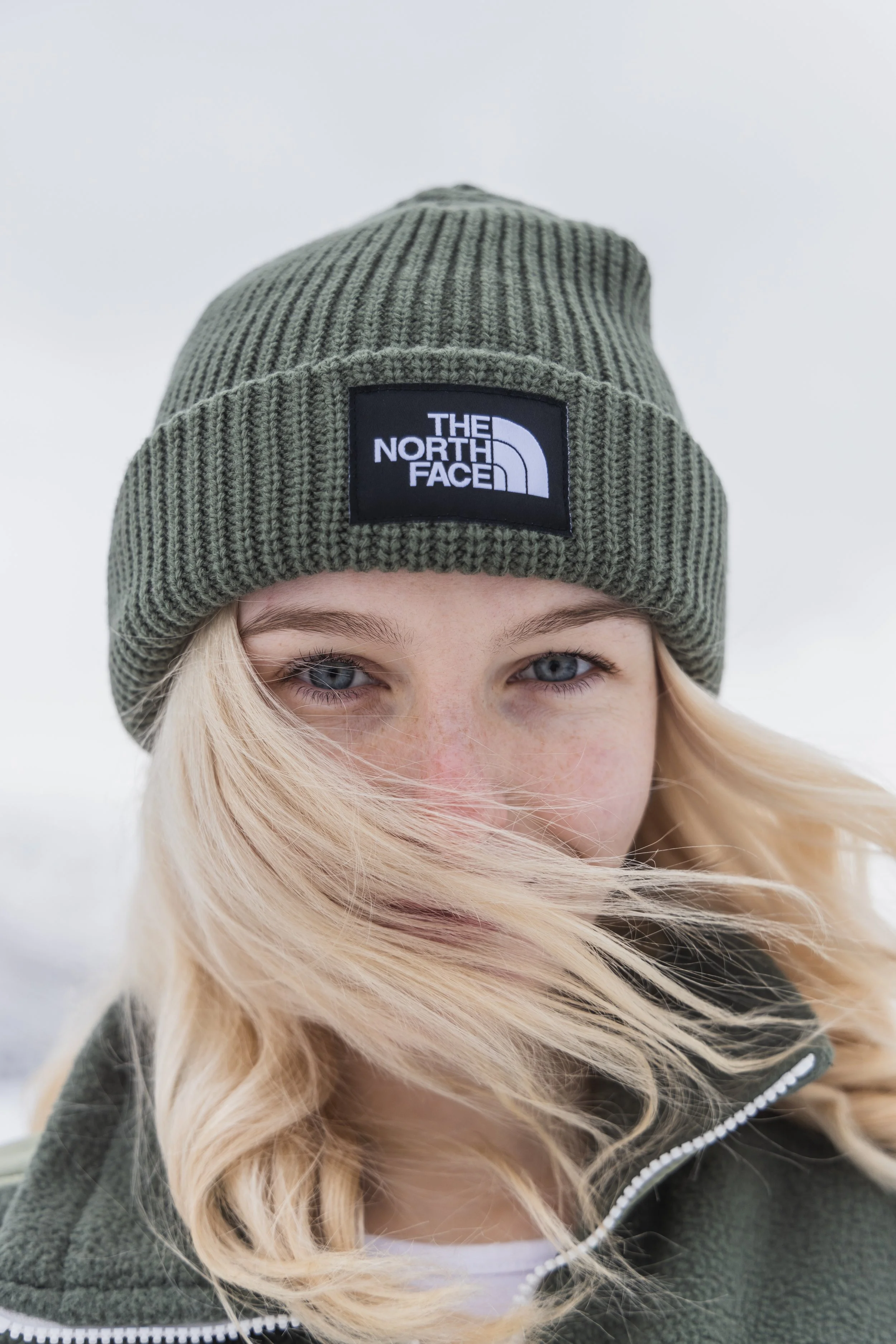 A young woman with blonde hair, wearing a green ribbed knit beanie with a black and white North Face logo patch, and a dark green zip-up jacket with a white shirt underneath, standing outdoors in snowy weather.