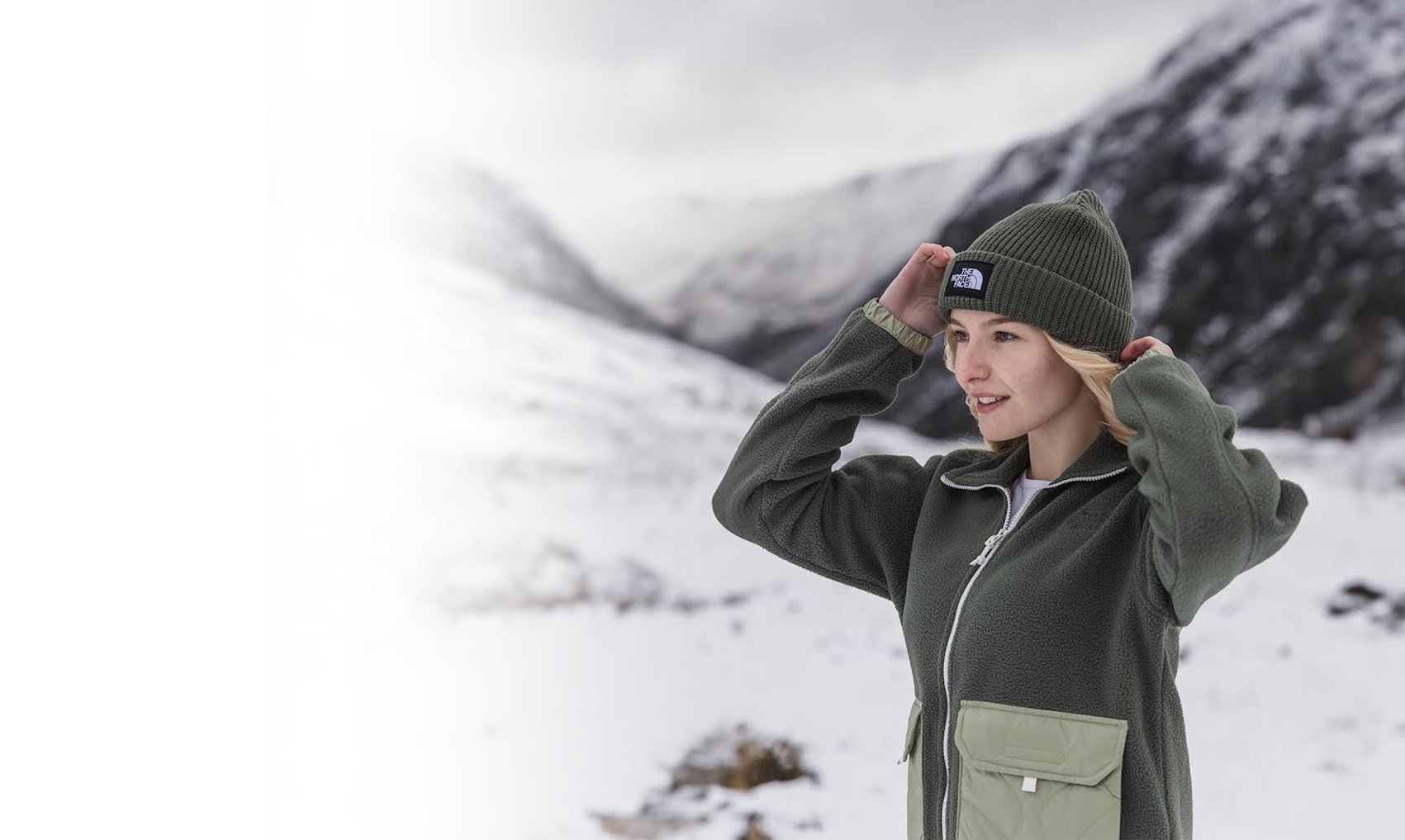 A woman wearing a green beanie and a gray jacket stands outdoors in snowy mountainous terrain, adjusting her hat and looking into the distance.