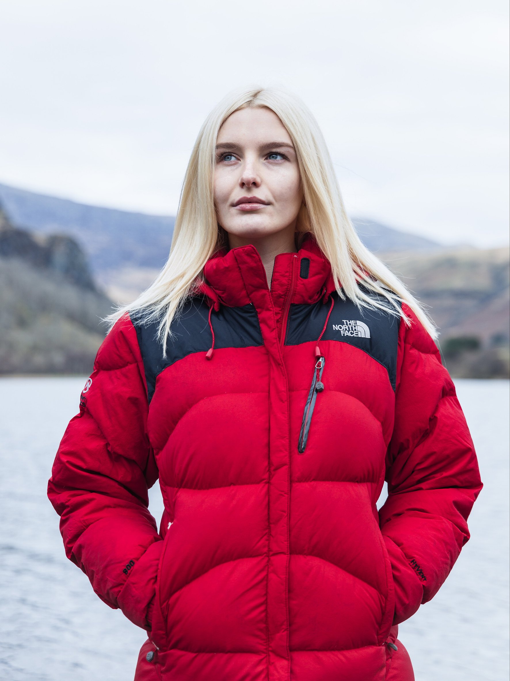 A young woman with blonde hair and blue eyes is wearing a red North Face winter jacket and standing outdoors near a body of water with mountains in the background, during overcast weather.