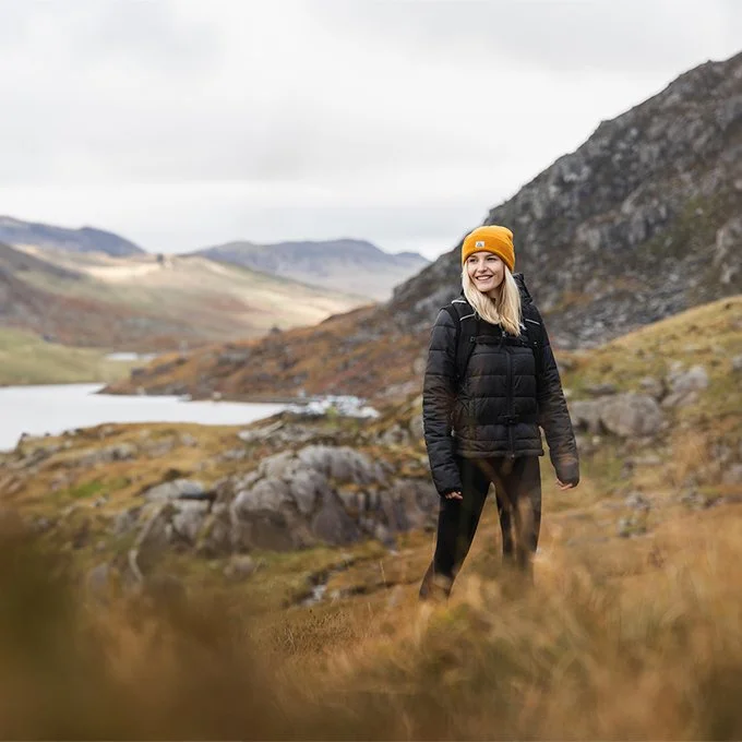 A woman standing outdoors on a hillside during daytime, wearing a black jacket, orange beanie, and backpack, with mountains, rocks, and a lake in the background.
