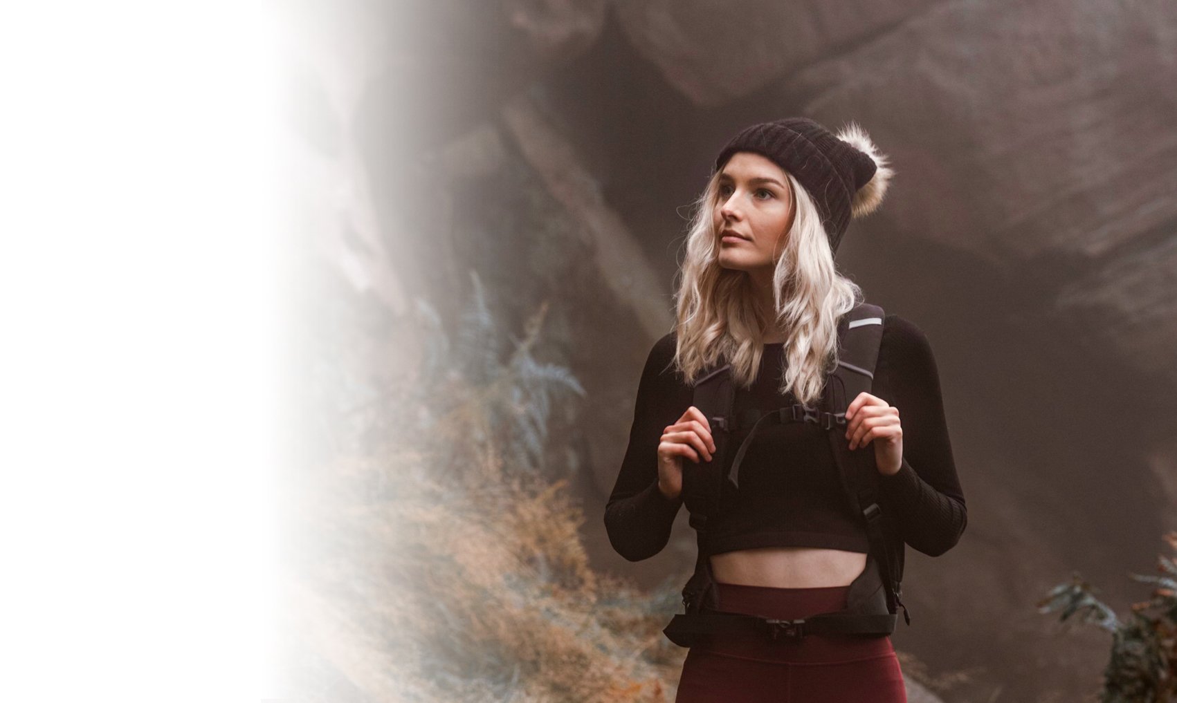 A woman with blonde hair wearing a black knit hat, black long-sleeve crop top, and burgundy leggings, carrying a backpack, standing outdoors near a rocky cliff or mountain in a natural setting.